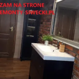 Umywalka z szafką w łazience z ceglaną ścianą, lustrem w srebrnej ramie i drewnianymi dodatkami, widoczny fragment drzwi i szafki, na ścianie napis z adresem strony internetowej.