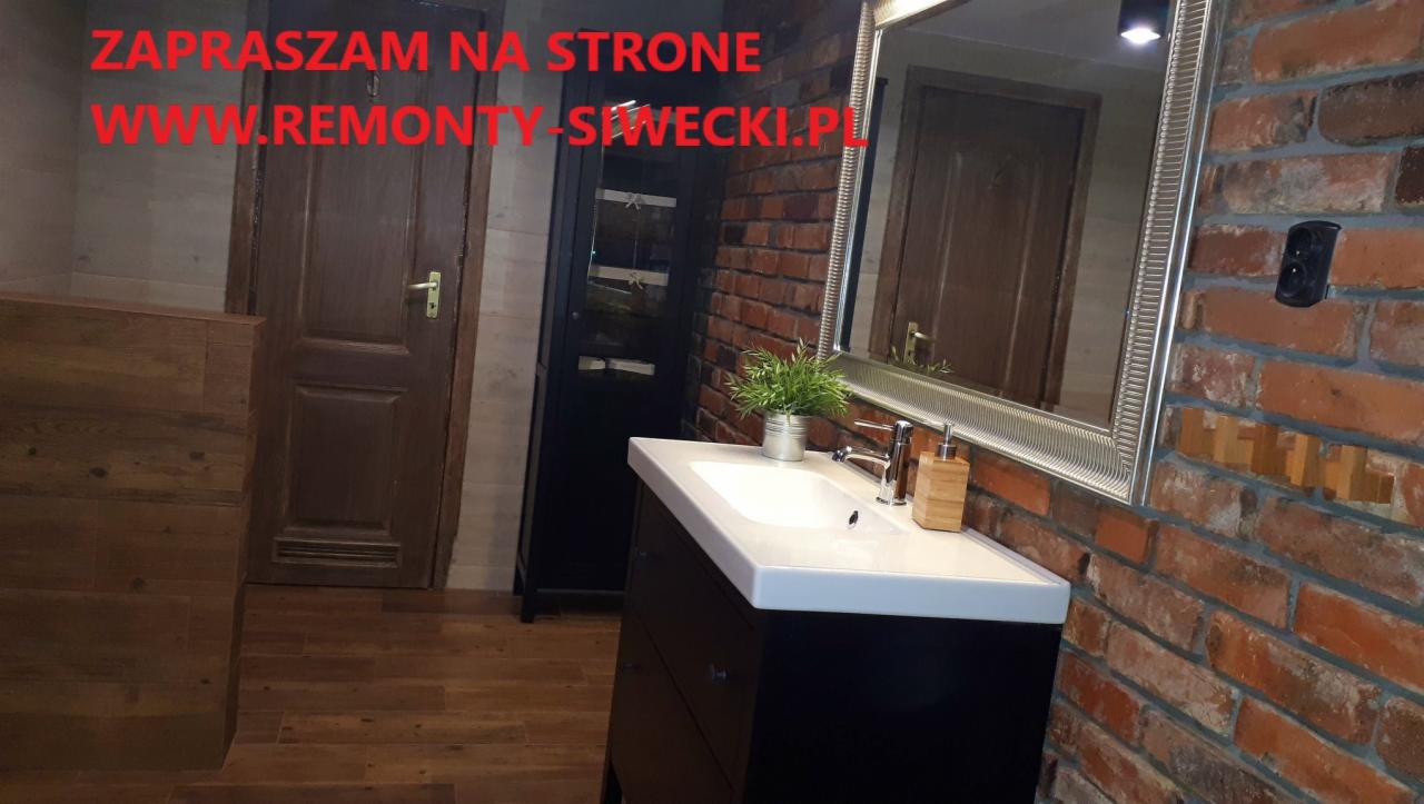 Umywalka z szafką w łazience z ceglaną ścianą, lustrem w srebrnej ramie i drewnianymi dodatkami, widoczny fragment drzwi i szafki, na ścianie napis z adresem strony internetowej.