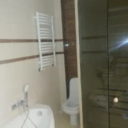 Nowoczesna łazienka z beżowymi płytkami, grzejnikiem drabinkowym, białą wanną z chromowaną armaturą i kabiną prysznicową z ciemnego szkła, obok toaleta.