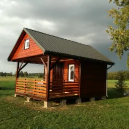 Drewniany domek letniskowy z zadaszonym tarasem, pomalowany na czerwono-brązowy kolor, z szarym dachem blaszanym, na fundamencie z betonowych bloczk&oacute;w, na zielonej łące pod zachmurzonym niebem.