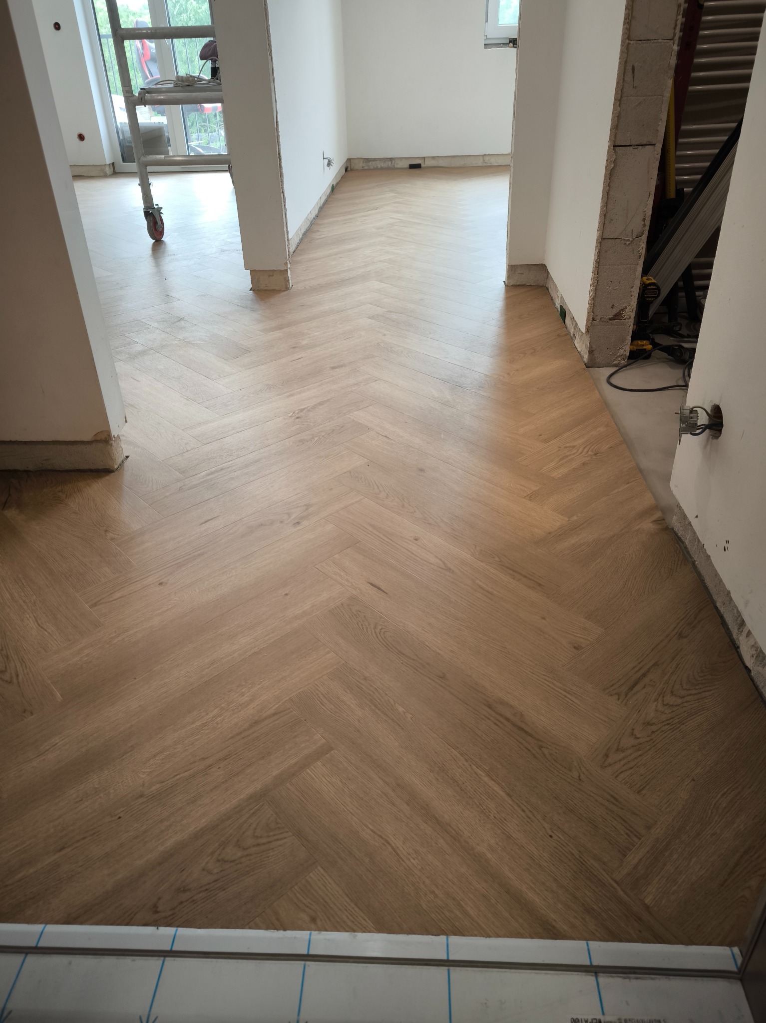 Nowo położona, jasna drewniana podłoga w jodełkę w trakcie remontu mieszkania, widoczne niedokończone ściany i drabina w tle.