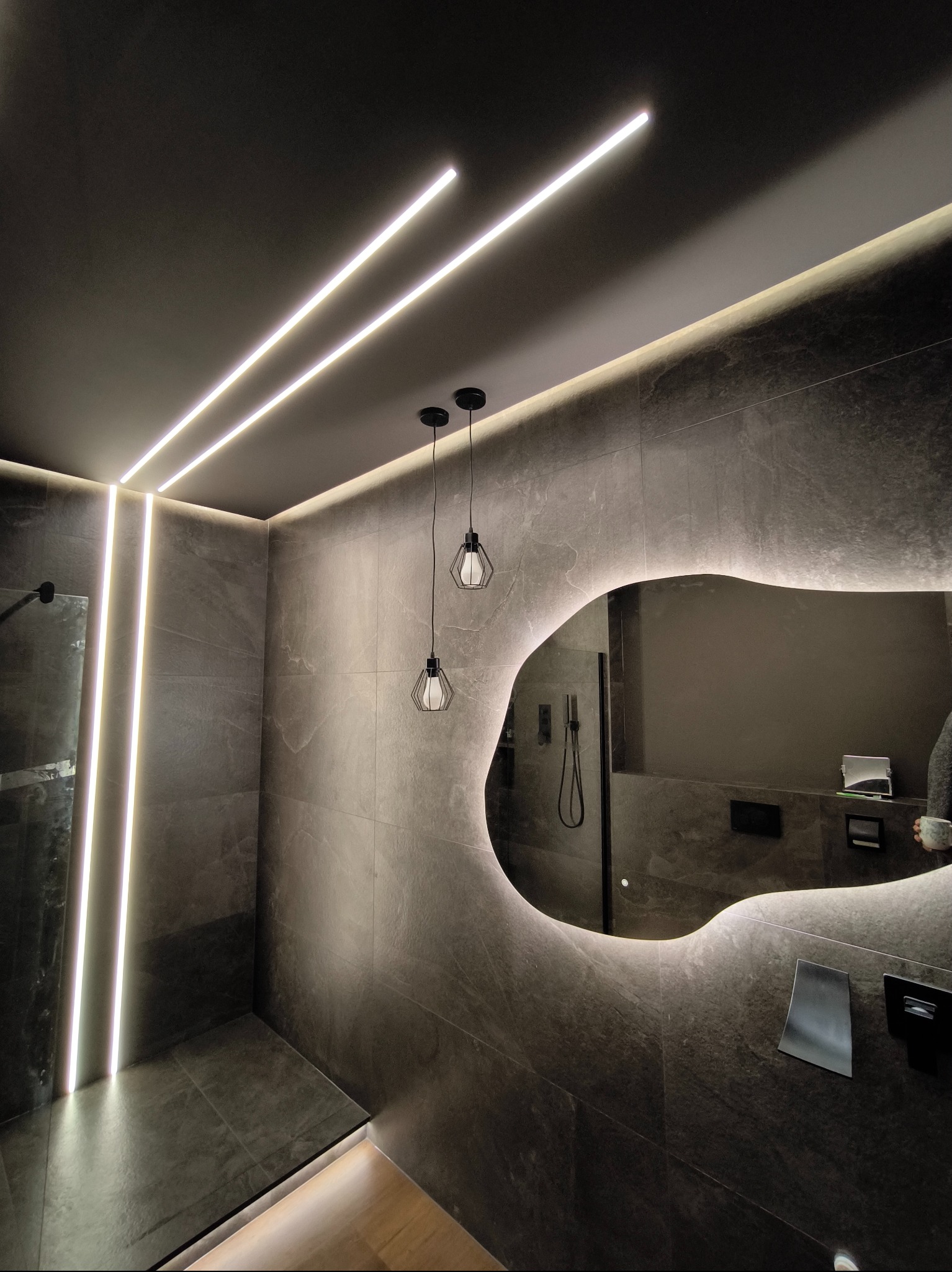 Nowoczesna łazienka z szarymi płytkami, podświetlanym lustrem o nieregularnym kształcie i liniowym oświetleniem LED na suficie, wiszące lampy w stylu industrialnym.