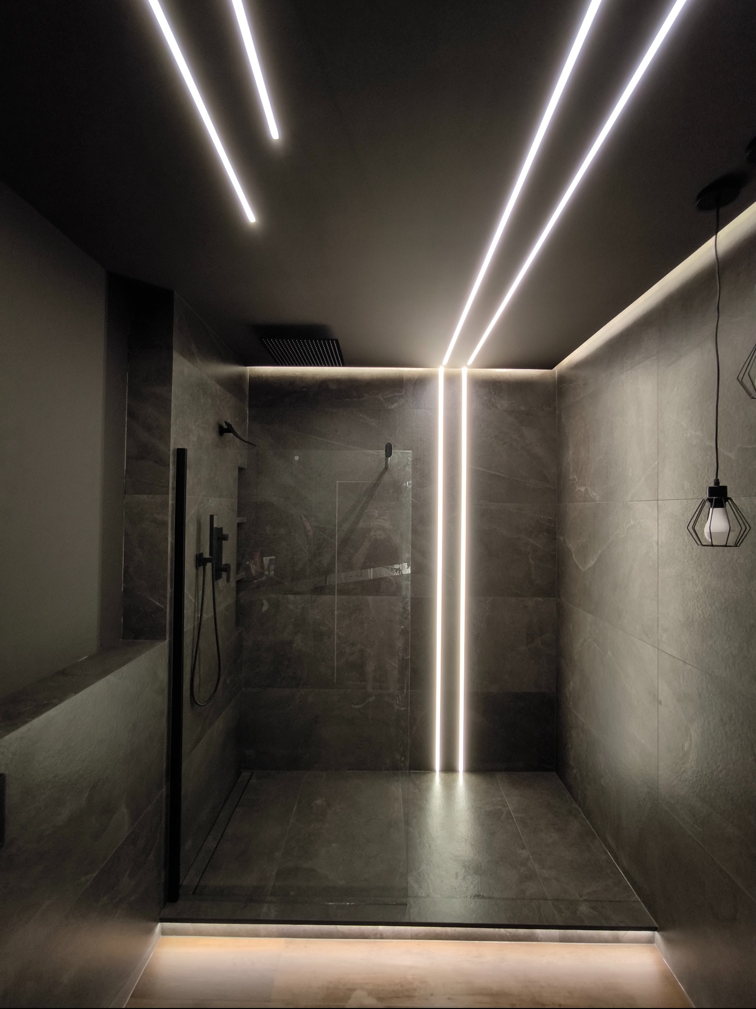 Nowoczesna kabina prysznicowa z szarymi płytkami, podświetleniem LED w suficie i wzdłuż ściany, czarną armaturą i minimalistyczną lampą wiszącą.