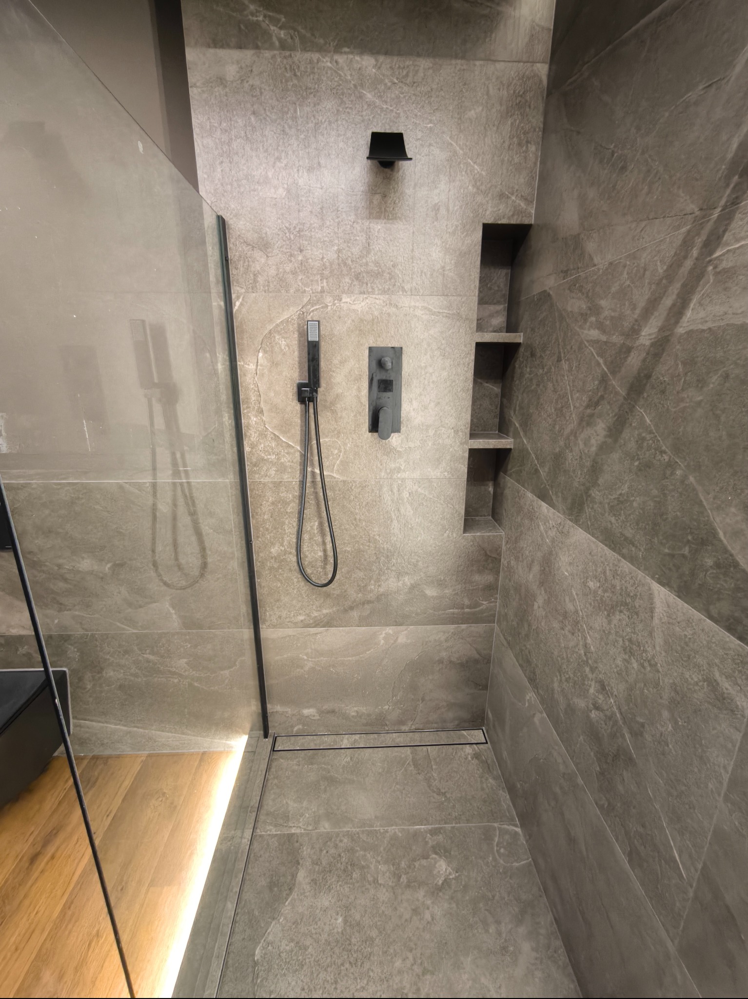 Nowoczesna kabina prysznicowa z szarymi płytkami imitującymi marmur, czarną armaturą i wnękami na kosmetyki, widoczne drewniane elementy podłogi poza kabiną.