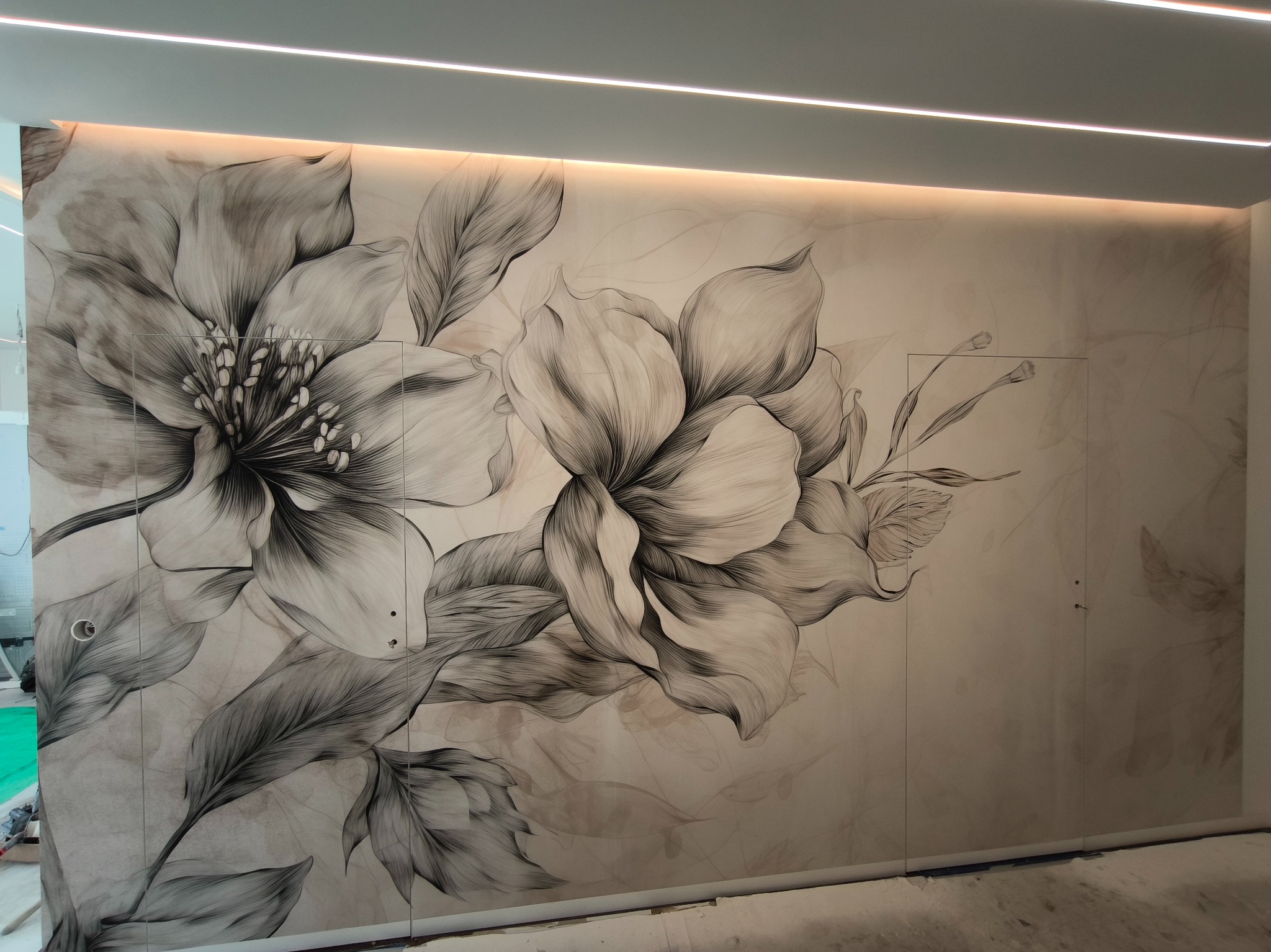 Ściana z dużym, ręcznie rysowanym motywem kwiatowym w odcieniach szarości, oświetlona od góry listwami LED, w trakcie prac wykończeniowych.