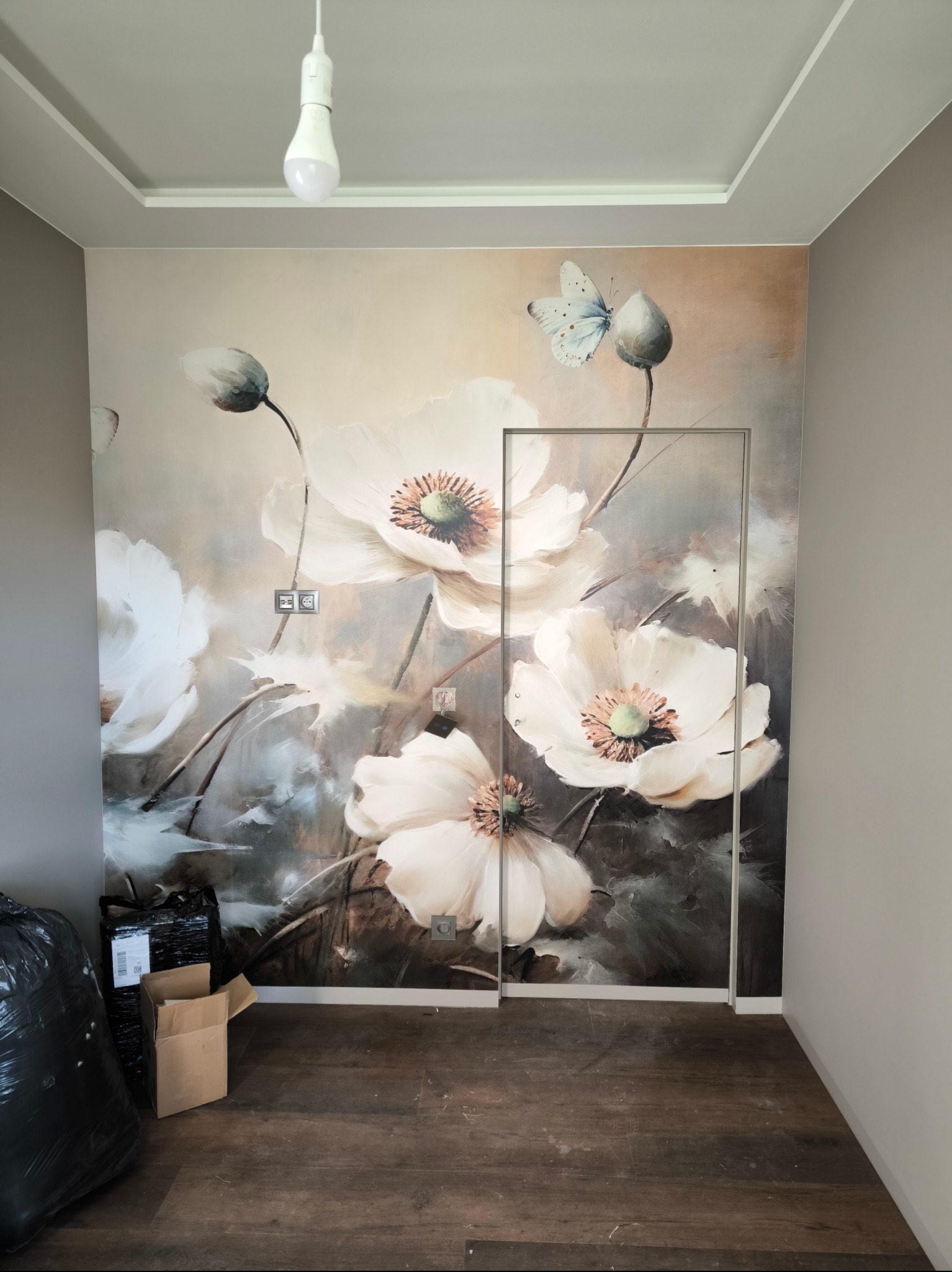 Pokój z fototapetą przedstawiającą białe kwiaty i motyla, widoczne gniazdka elektryczne na tapecie, częściowo zamontowana ościeżnica drzwiowa, podłoga z paneli, worki z odpadami budowlanymi w rogu.