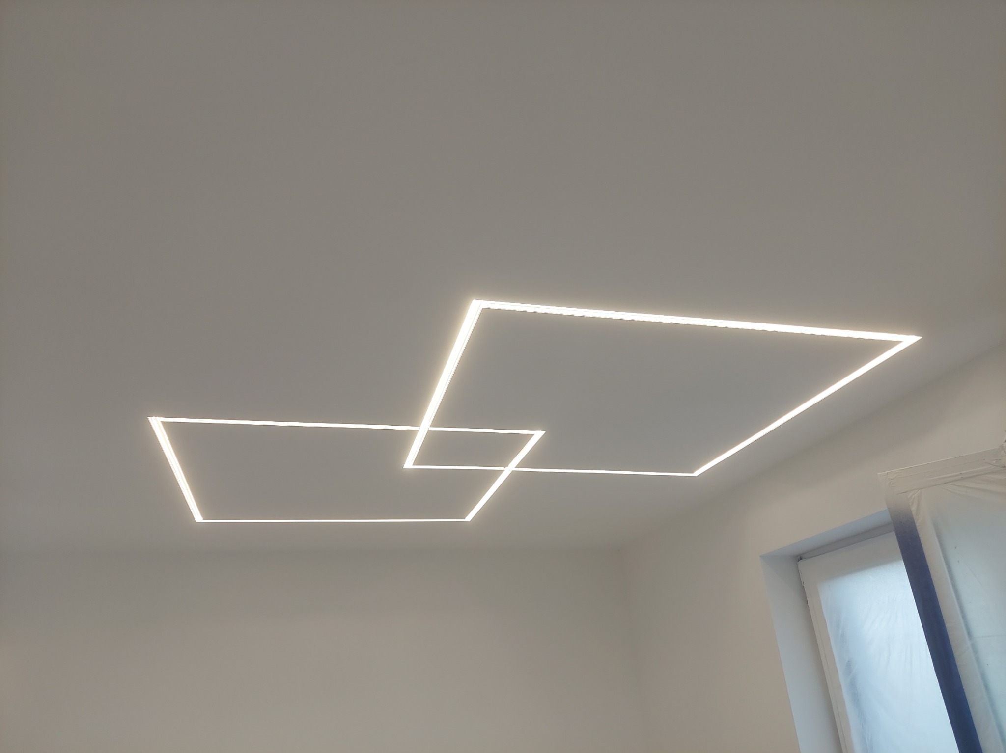 Nowoczesne oświetlenie sufitowe: Dwa przecinające się kwadraty LED w minimalistycznym wnętrzu, widoczne okno zabezpieczone folią malarską.