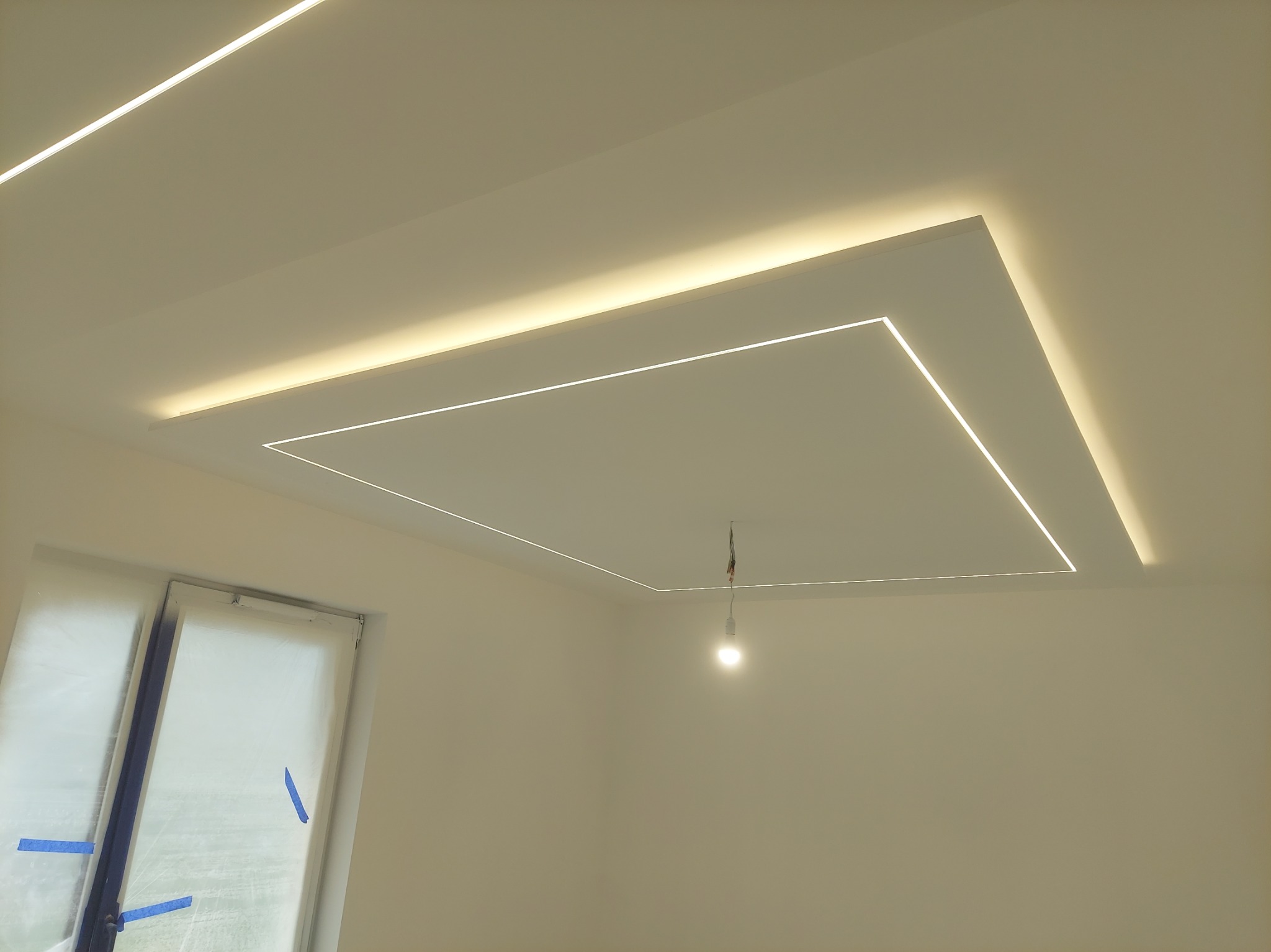 Nowoczesny sufit podwieszany z oświetleniem LED w kształcie kwadratu, widoczne okno zabezpieczone folią malarską, żarówka tymczasowa na kablu.