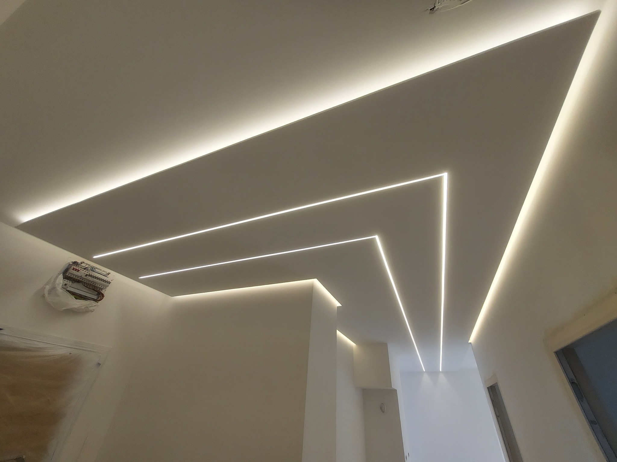 Nowoczesne oświetlenie sufitowe z liniowymi oprawami LED tworzącymi geometryczny wzór w białym wnętrzu, widoczna niezabezpieczona instalacja elektryczna.