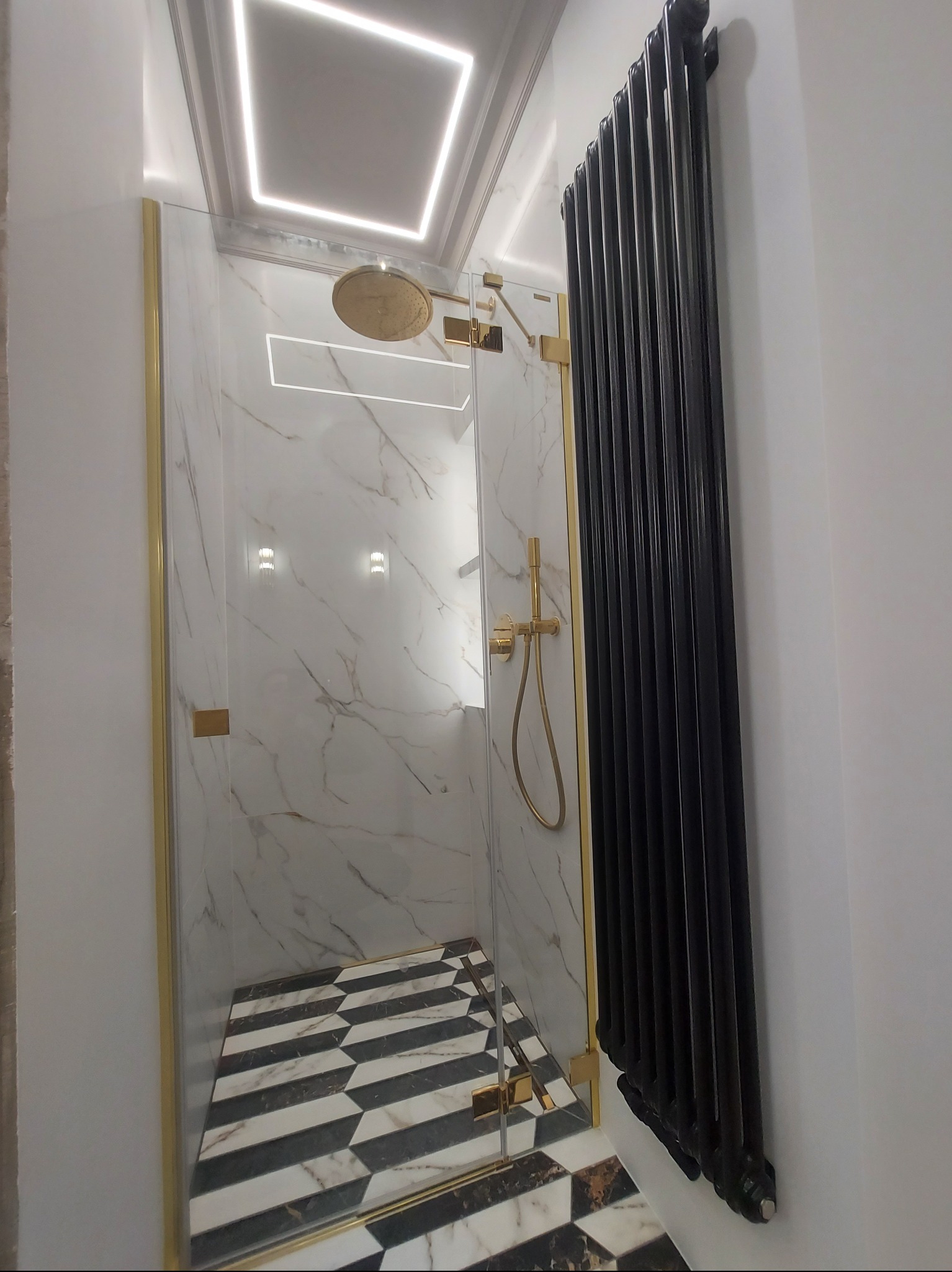 Nowoczesna kabina prysznicowa z marmurowymi ścianami, złotymi detalami i czarno-białą podłogą w jodełkę, obok grzejnik żeberkowy.