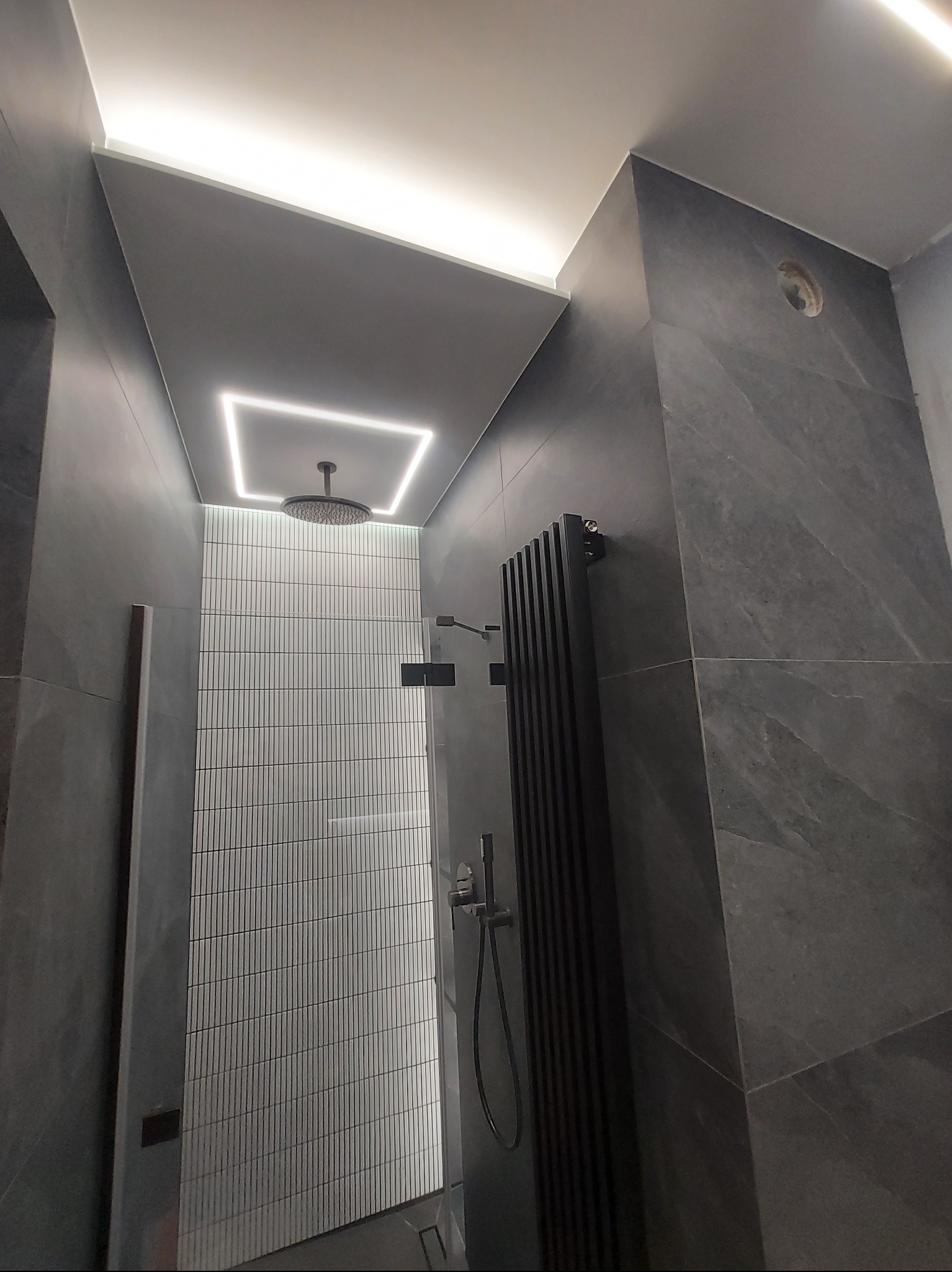 Nowoczesna łazienka z szarymi płytkami imitującymi kamień, podświetlanym sufitem i deszczownicą.