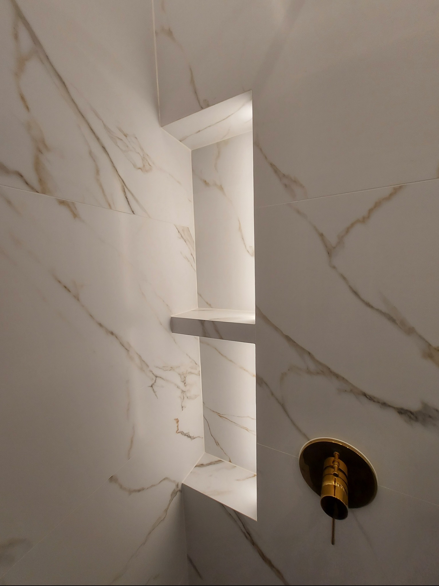 Narożna wnęka z półkami w kabinie prysznicowej wyłożonej białymi płytkami imitującymi marmur z złotymi żyłami, widoczny złoty zawór.