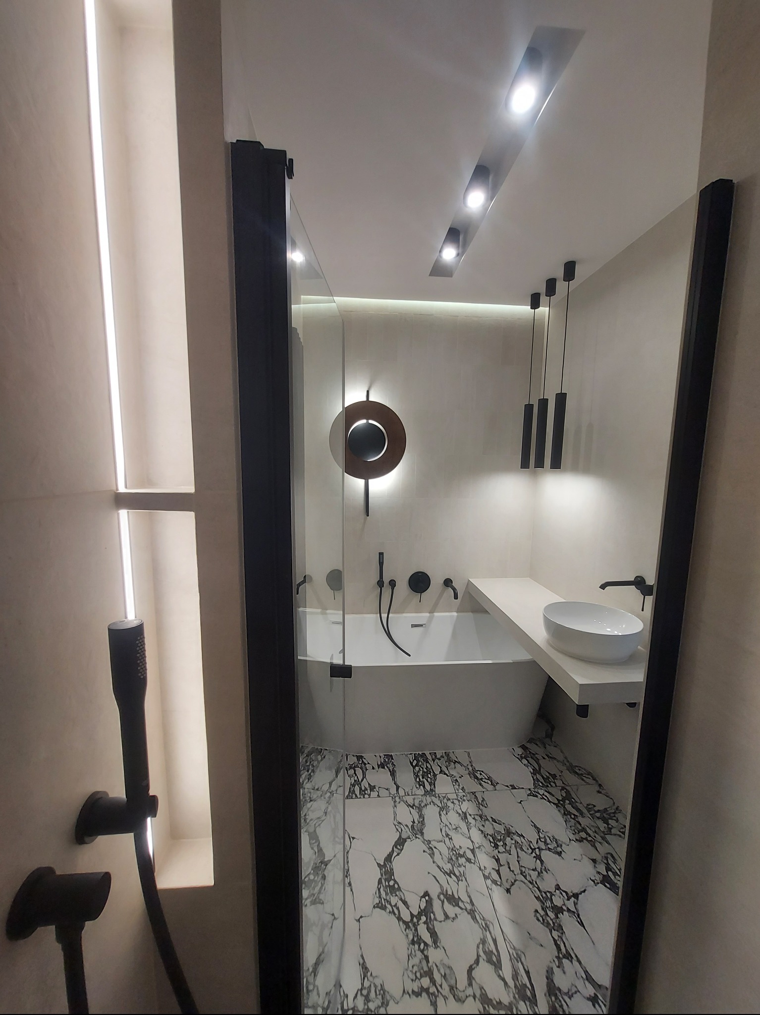 Nowoczesna łazienka z wanną, umywalką nablatową, czarną armaturą i marmurową podłogą, oświetlona ledowymi lampami sufitowymi.