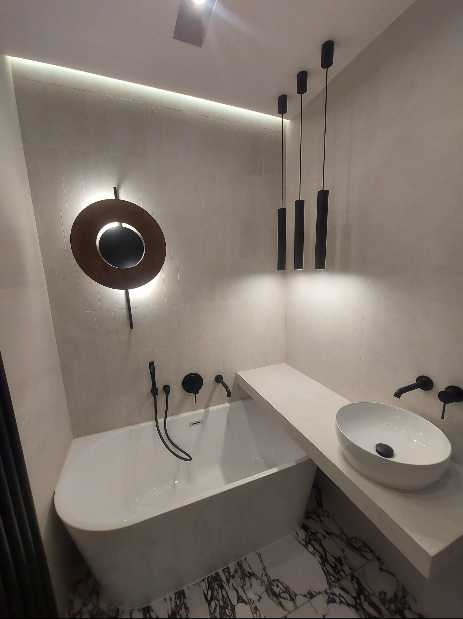 Nowoczesna łazienka z owalną wanną, umywalką nablatową i oryginalnym oświetleniem: okrągła lampa ścienna i trzy cylindryczne lampy wiszące, wykończona w jasnych kolorach z akcentami czerni.