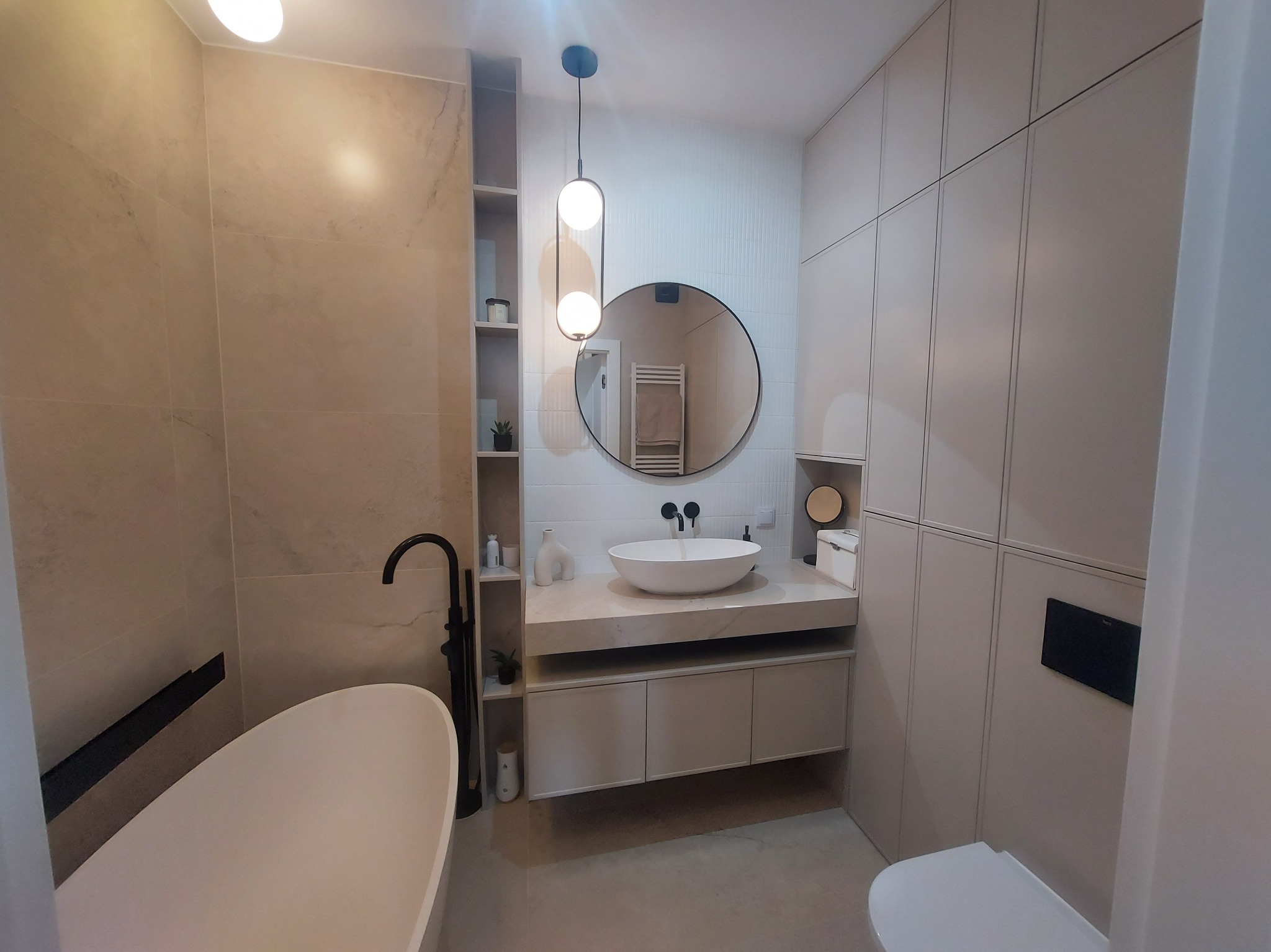 Nowoczesna łazienka z wolnostojącą wanną, okrągłym lustrem, wiszącą lampą z dwoma kulami i szafką z umywalką nablatową, utrzymana w jasnej kolorystyce.