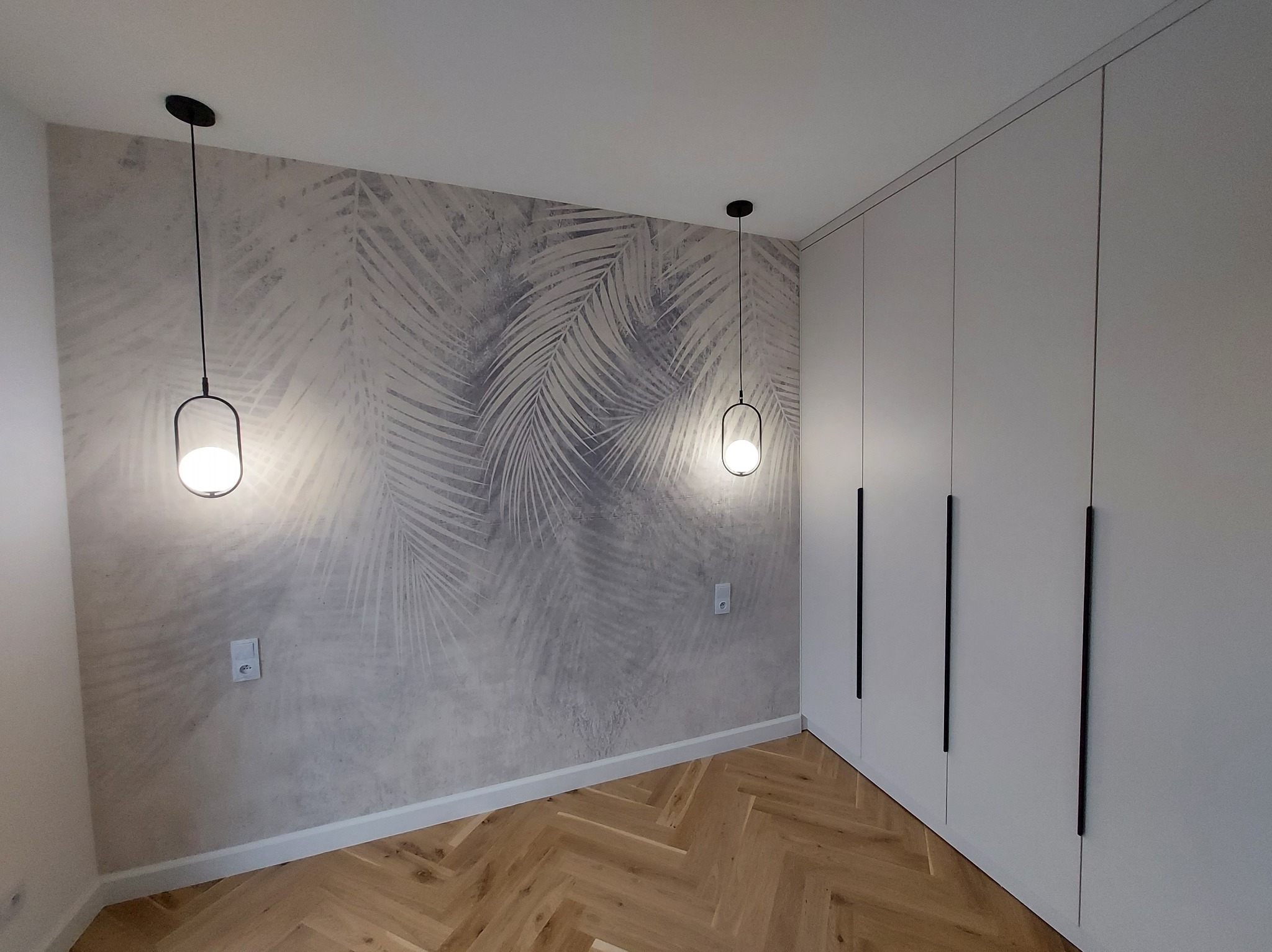 Nowoczesna sypialnia z drewnianą podłogą w jodełkę, tapetą w motywy liści palmowych, minimalistycznymi lampami wiszącymi i białą zabudową szafy z długimi, czarnymi uchwytami.
