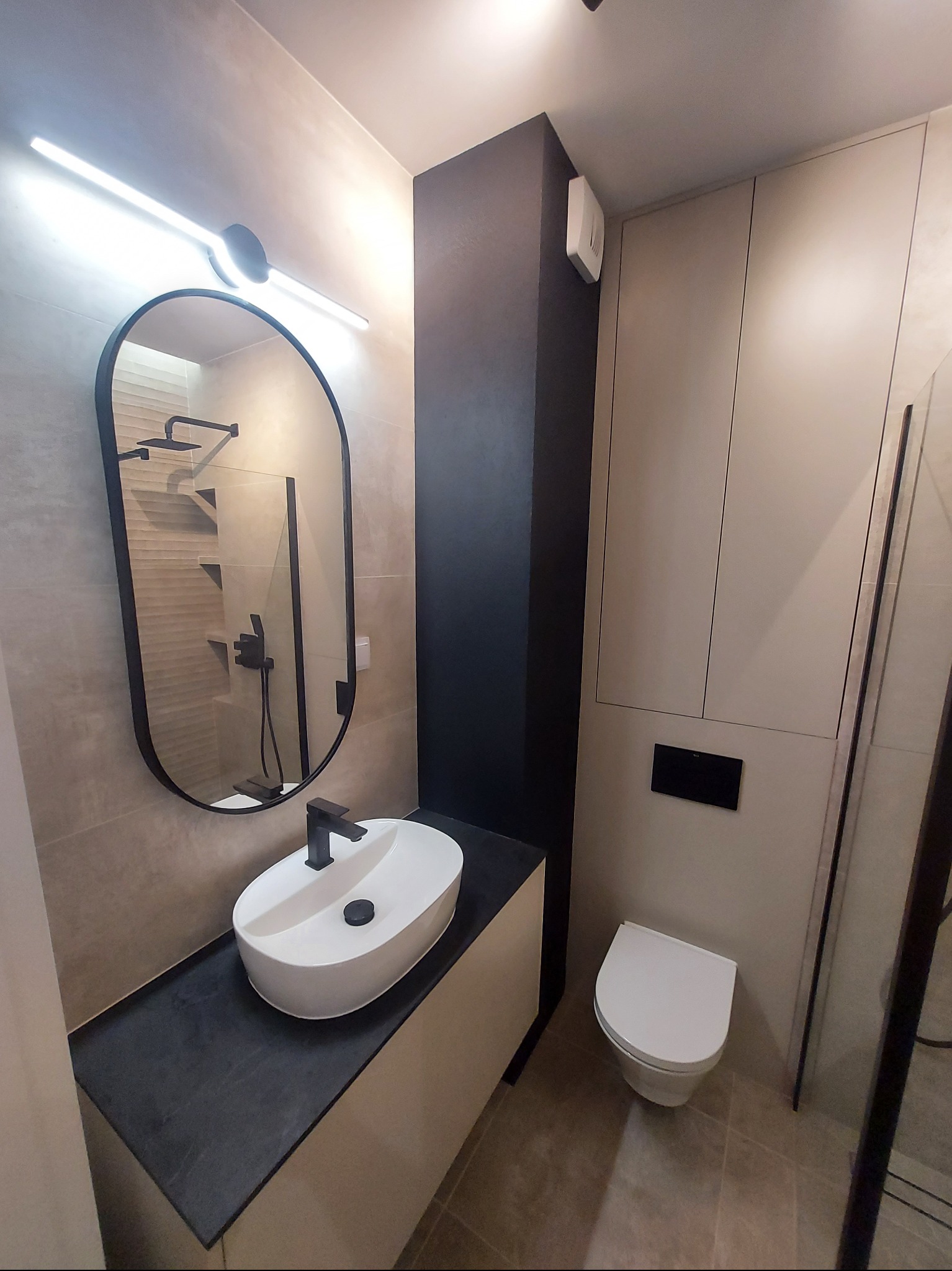 Nowoczesna łazienka z owalną umywalką nablatową na czarnym blacie, podświetlanym lustrem w czarnej ramie i minimalistyczną toaletą.