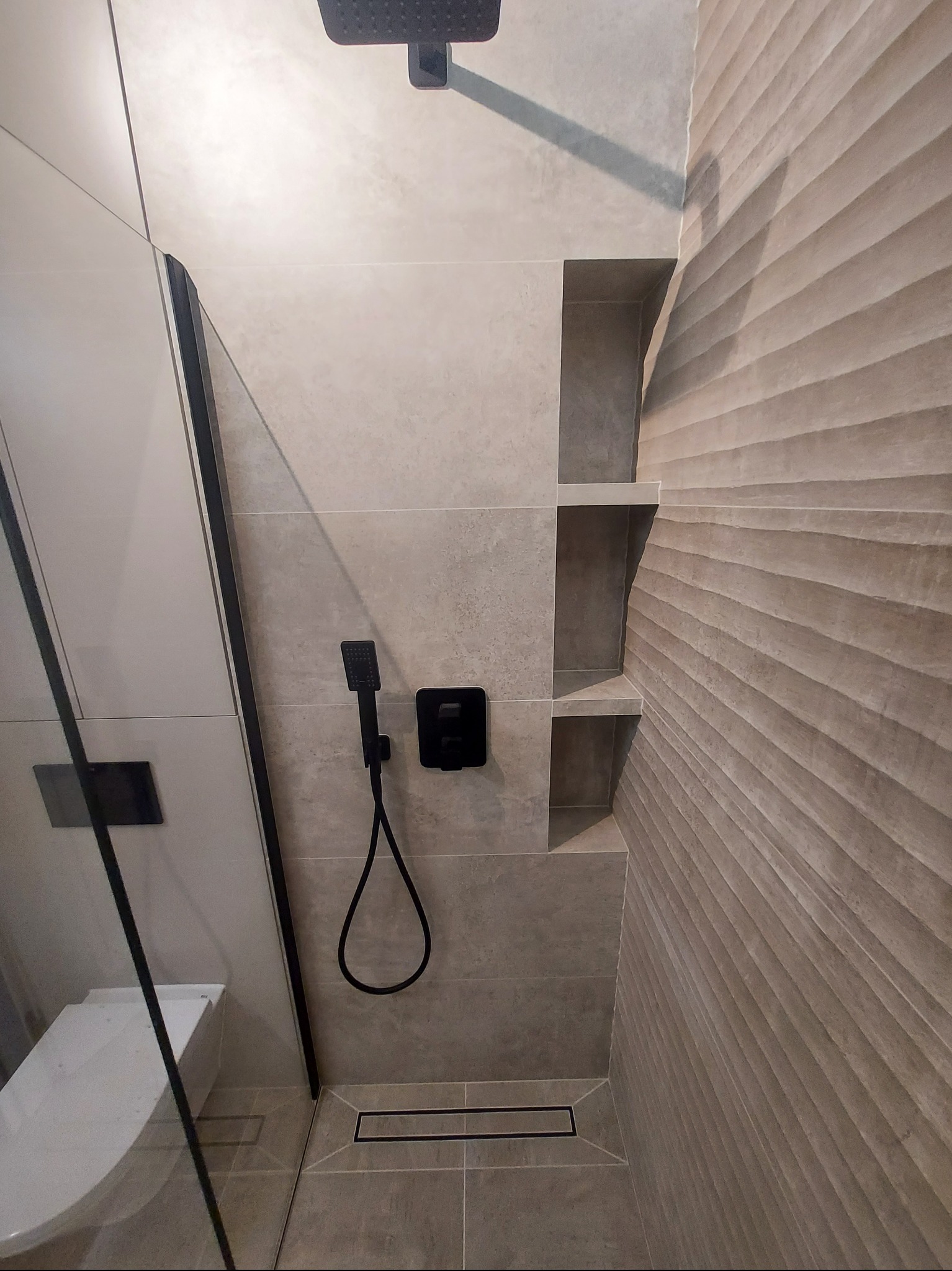 Nowoczesna łazienka z kabiną prysznicową typu walk-in, czarną armaturą i półkami w ścianie wyłożonej płytkami o falistej strukturze. Widoczny odpływ liniowy w podłodze.