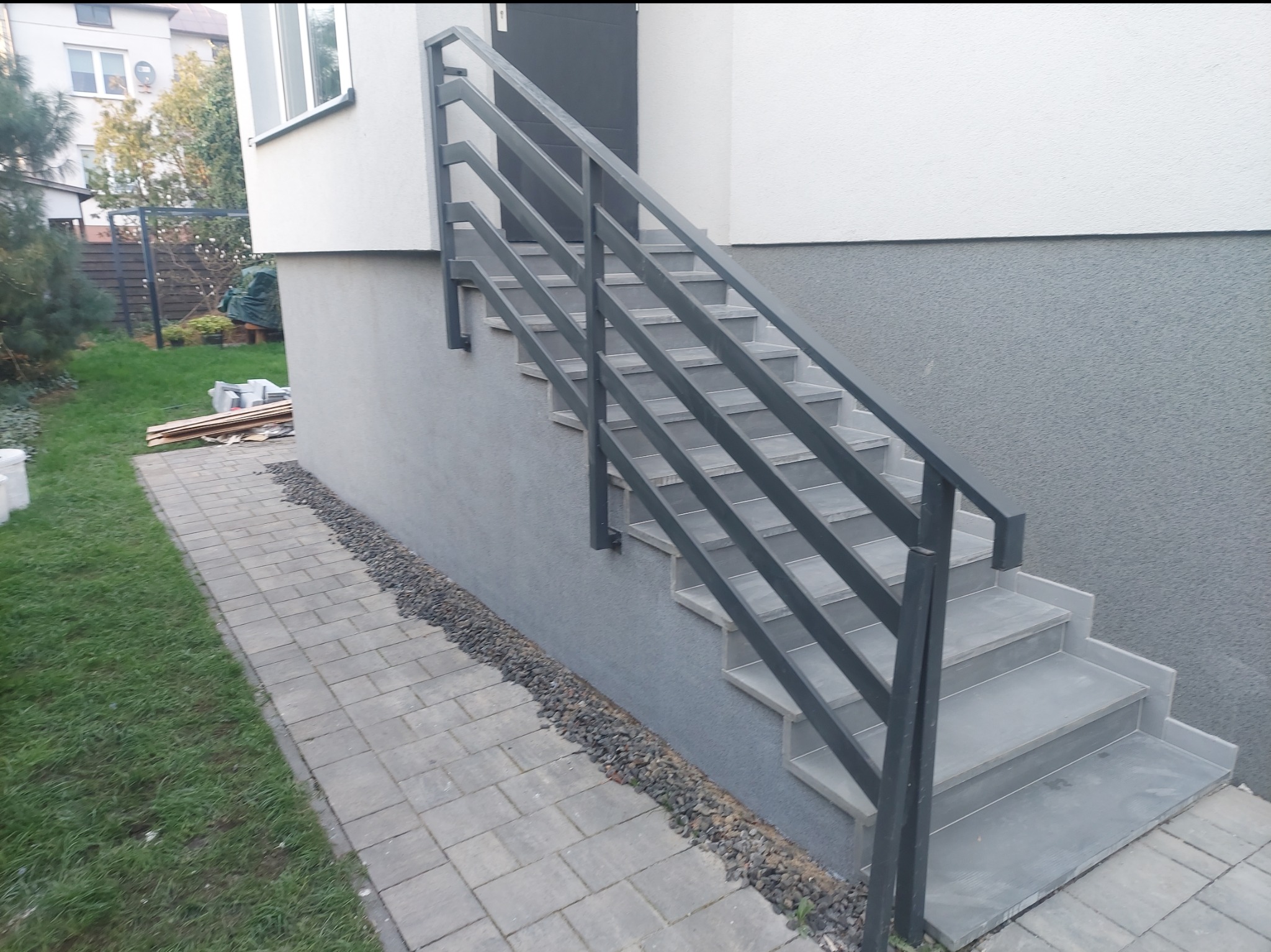 Zewnętrzne schody z szarą metalową balustradą prowadzące do wejścia domu, wykończone szarą kostką brukową i kamieniami ozdobnymi.