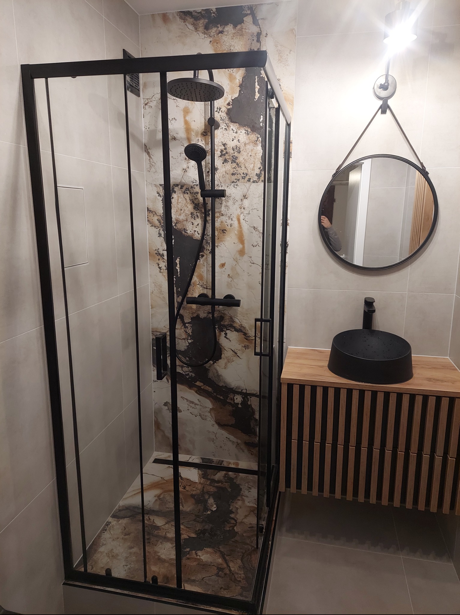 Nowoczesna łazienka z czarną kabiną prysznicową typu walk-in, umywalką nablatową w kolorze czarnym i okrągłym lustrem na tle beżowych płytek.