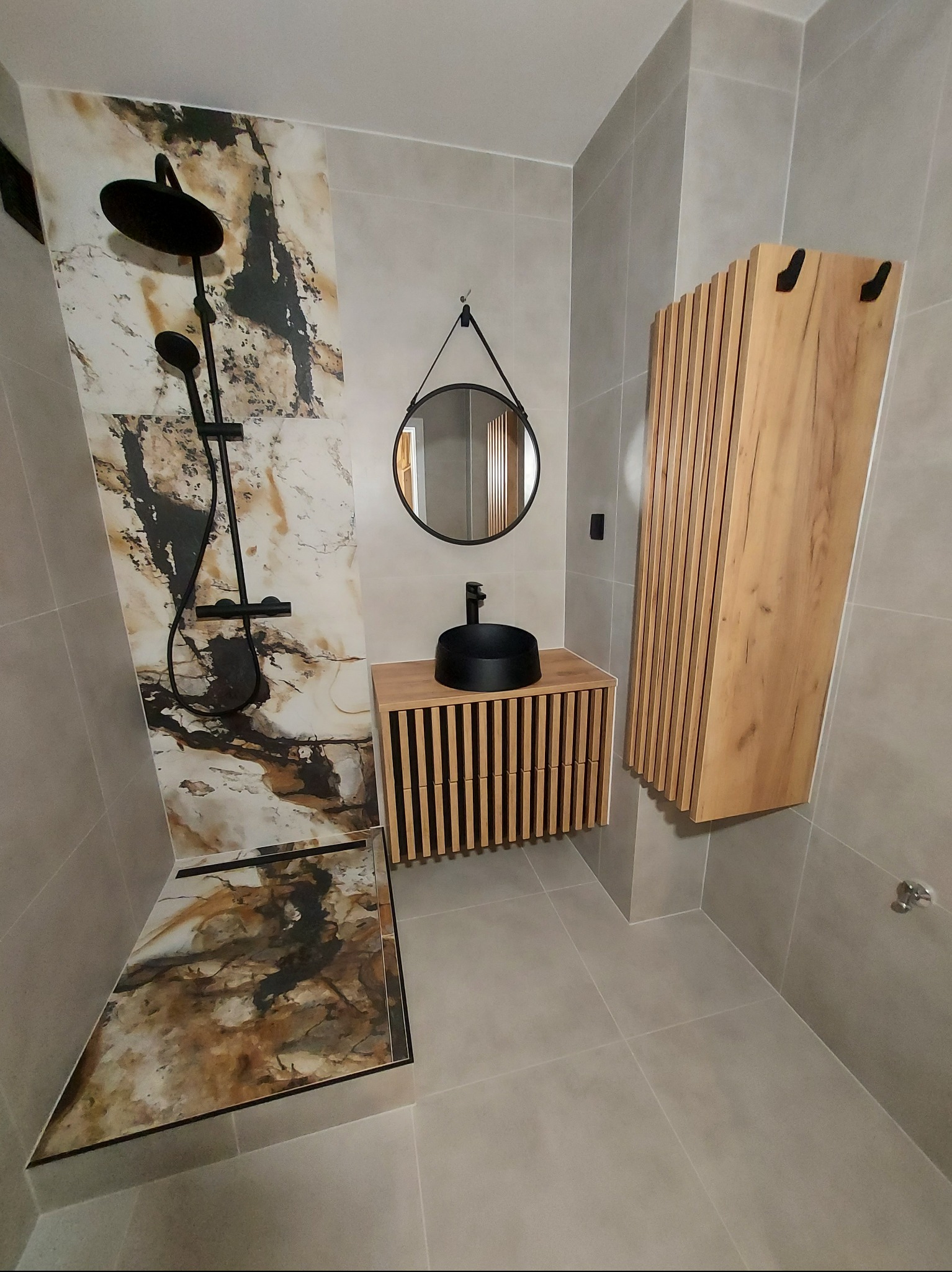 Nowoczesna łazienka z kabiną prysznicową wykończoną płytami imitującymi marmur, czarną armaturą, okrągłym lustrem i drewnianą szafką z lamelami.