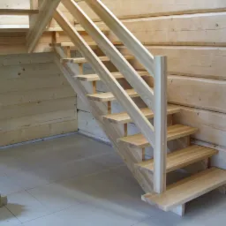 Jasne, drewniane schody z balustradą w domu z bali, widoczne fragmenty osoby w jeansach i narzędzia.