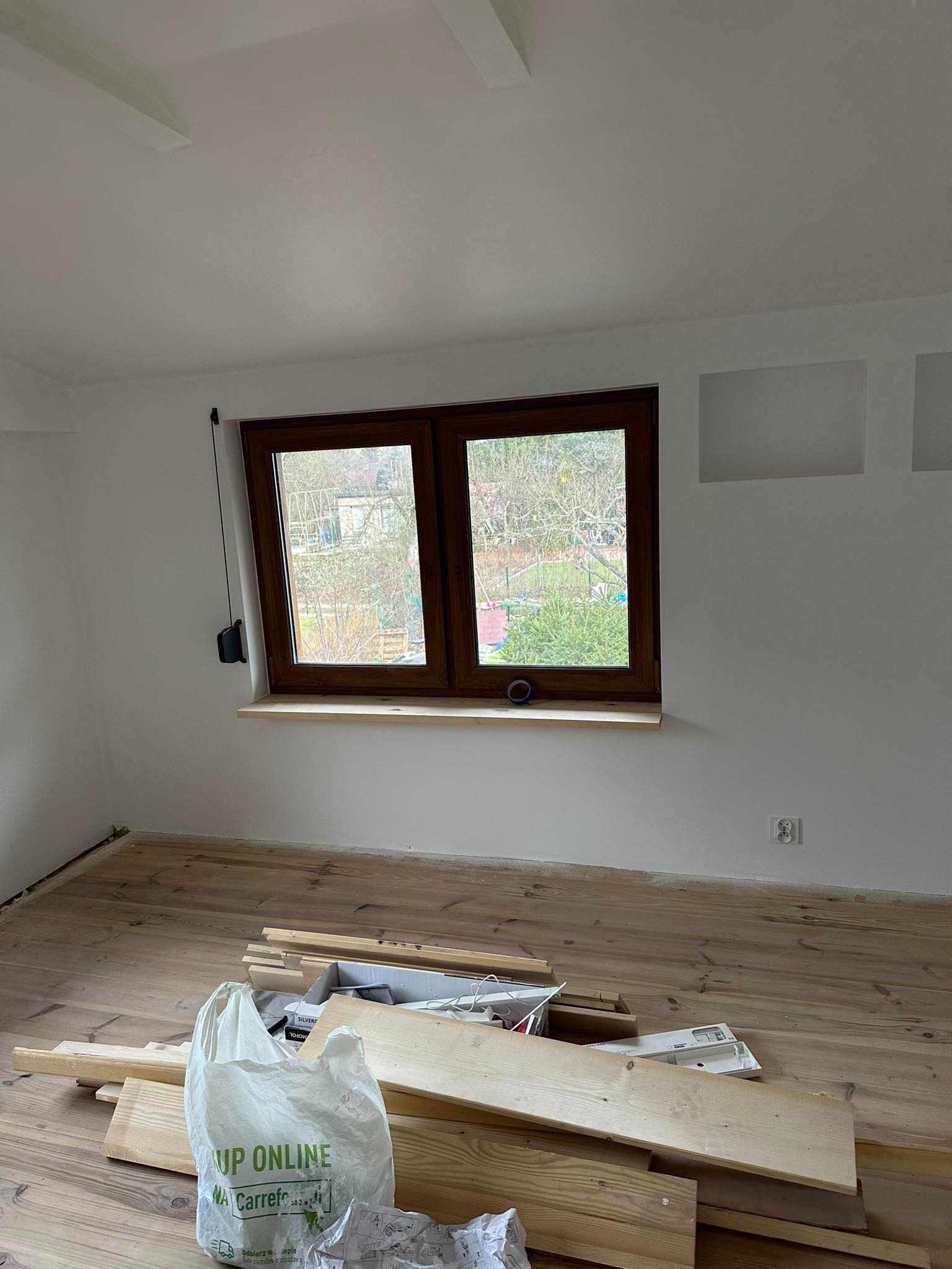 Pokój w trakcie prac wykończeniowych: świeżo pomalowane ściany, drewniana podłoga, okno z drewnianą ramą i przygotowane deski na podłodze.