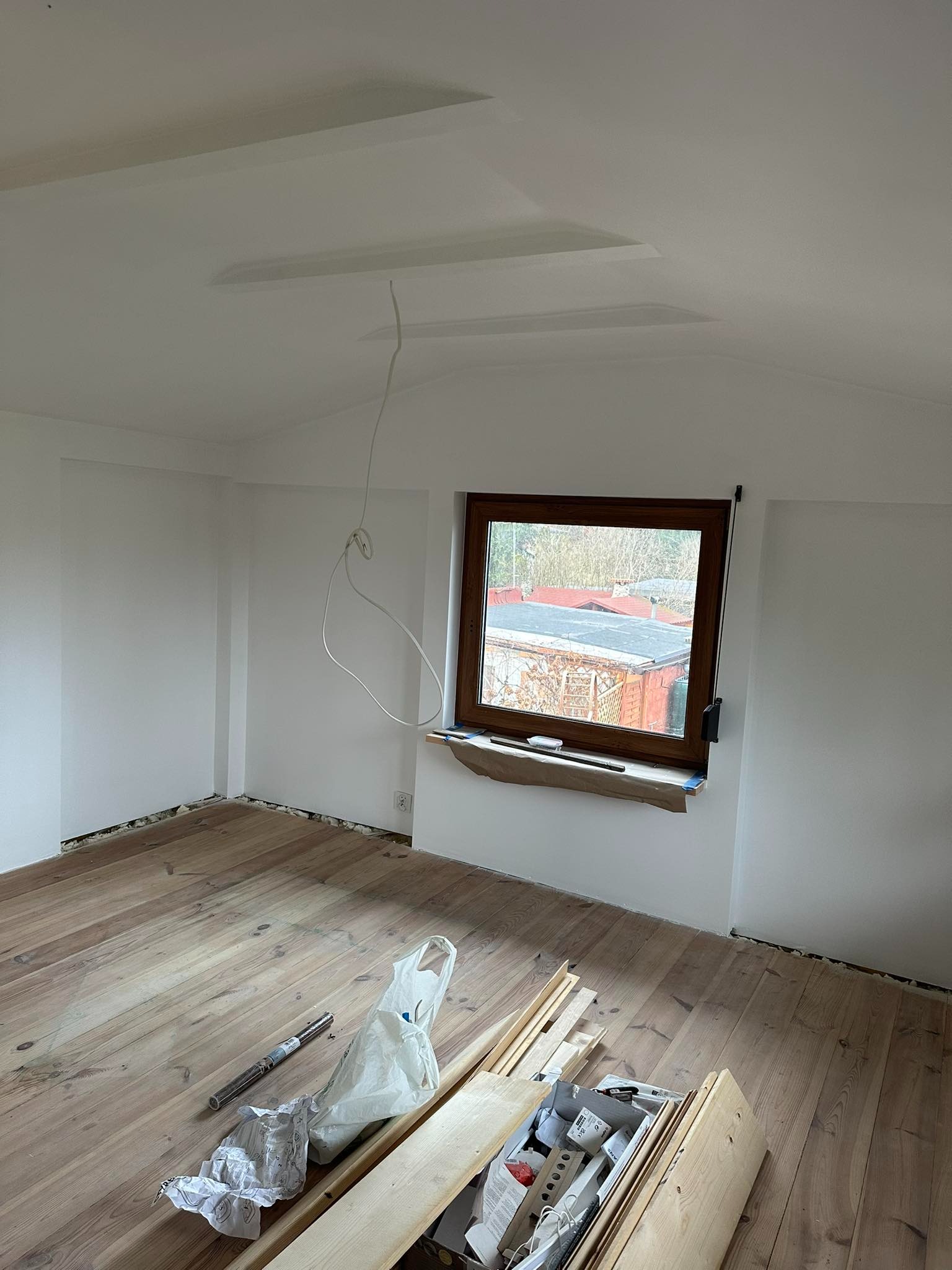 Wnętrze pokoju w trakcie remontu: świeżo pomalowane białe ściany i sufit z wystającymi kablami, drewniane okno z widokiem na zewnątrz, zabezpieczone folią, oraz drewniana podłoga z narzędziami...