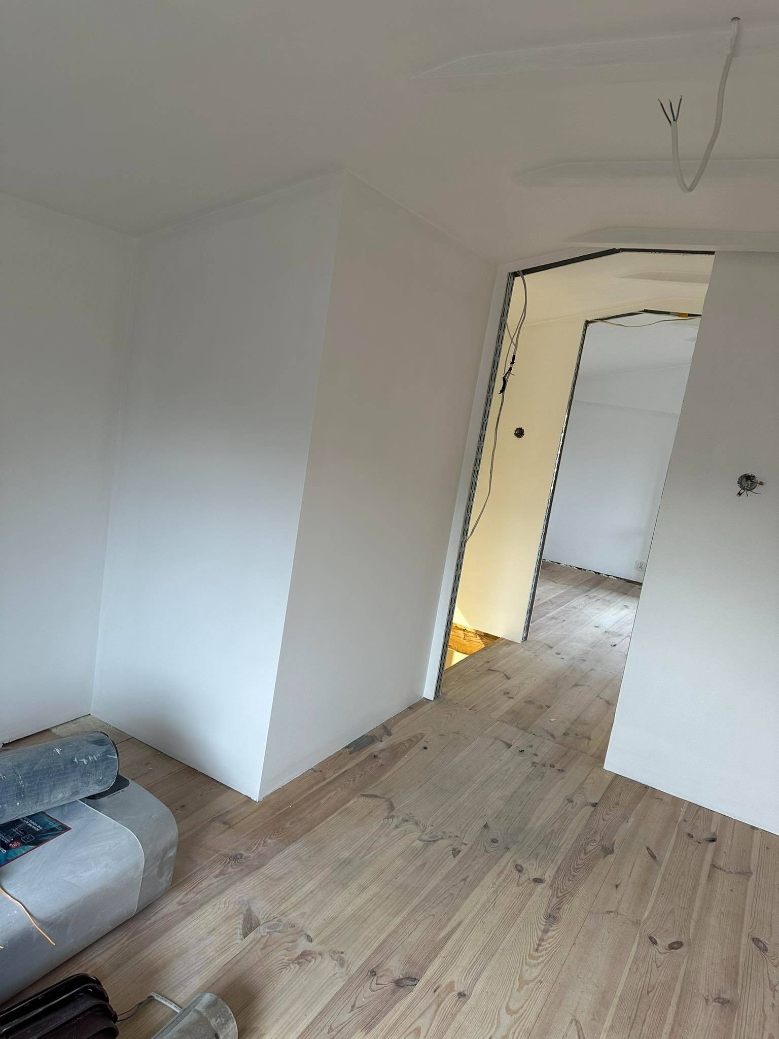 Wnętrze w trakcie prac wykończeniowych: świeżo pomalowane na biało ściany, drewniana podłoga, widoczne przewody elektryczne i narzędzia malarskie przy ścianie.