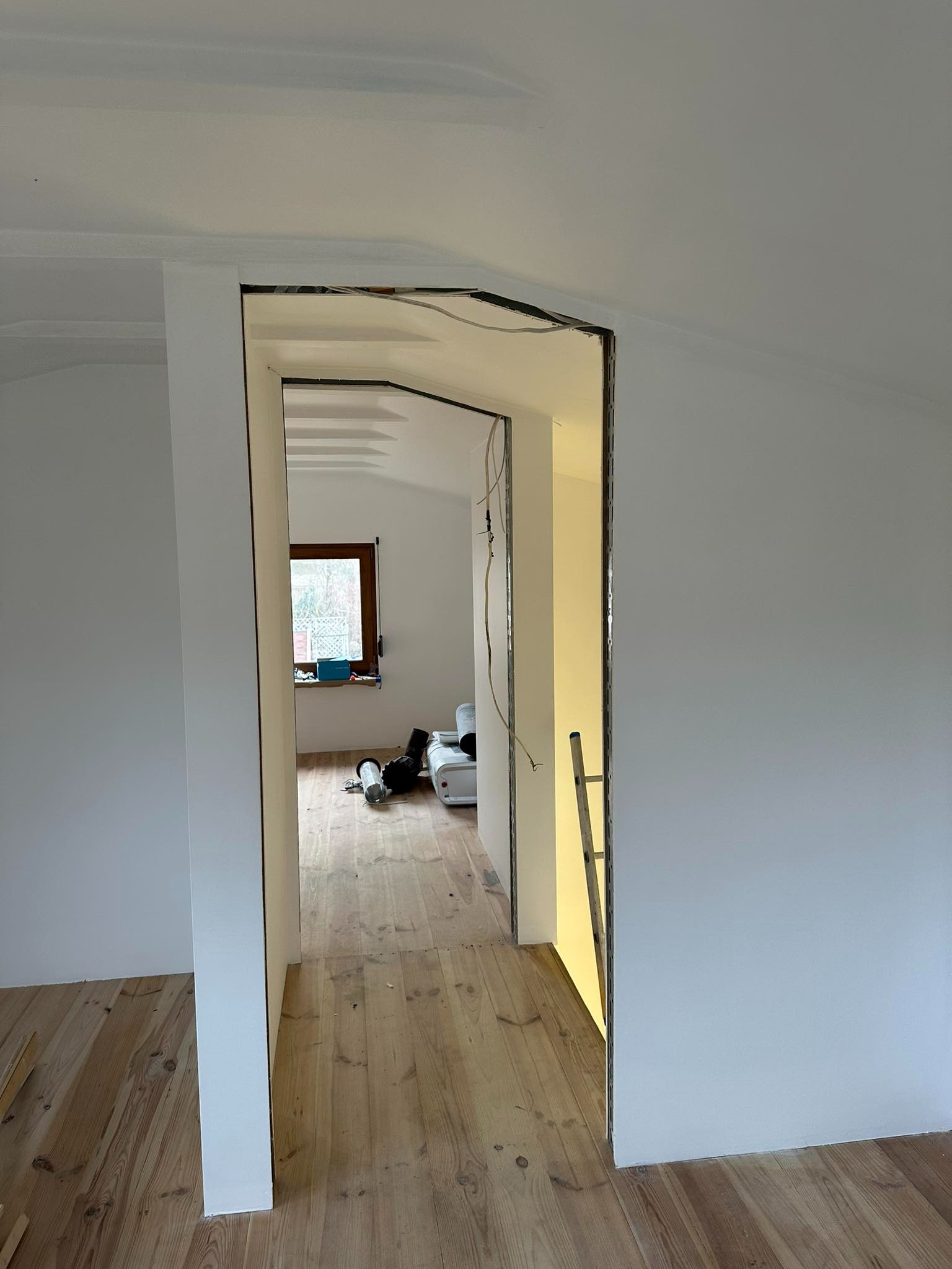 Wnętrze poddasza w trakcie prac: widok przez otwór w ścianie z widocznymi elementami konstrukcyjnymi, drewnianą podłogą i oknem w tle. Na podłodze leżą narzędzia i materiały budowlane.