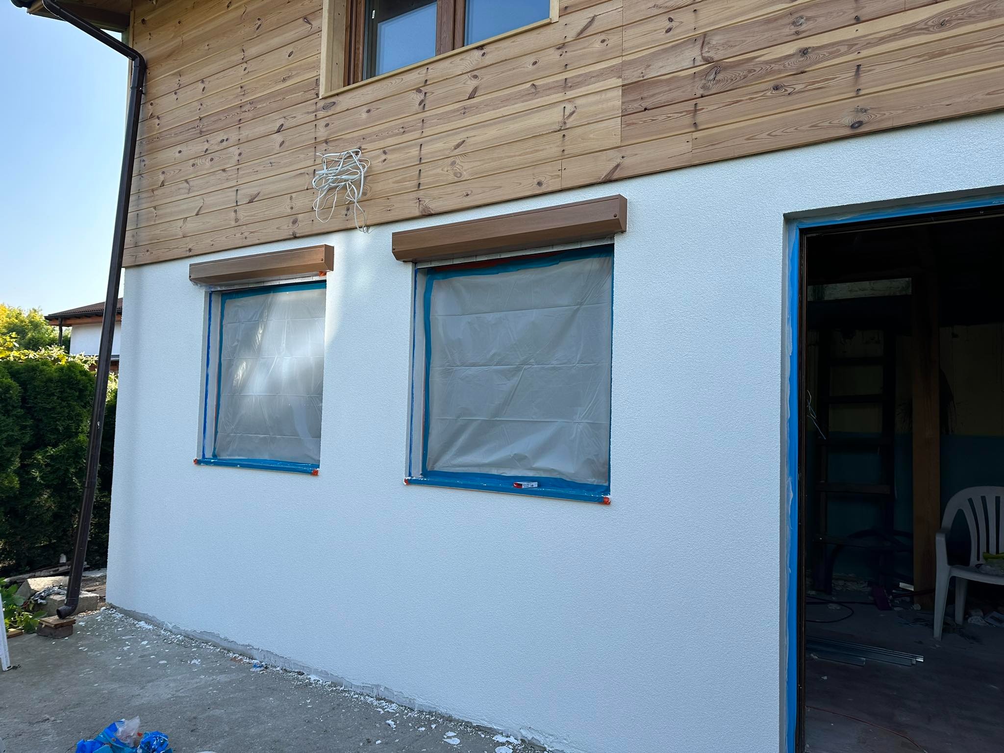 Elewacja domu z drewnianą boazerią i świeżo nałożoną, białą strukturą tynku, okna zabezpieczone folią malarską, widoczne ślady prac na podłożu.