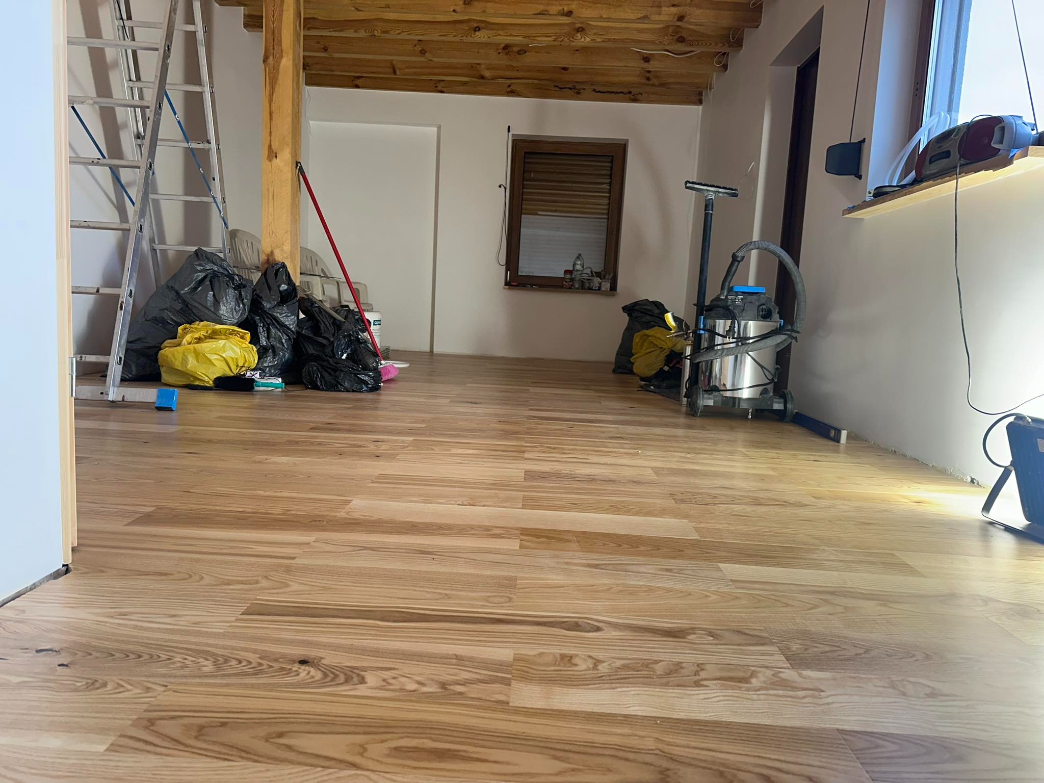 Wykończona drewniana podłoga w pomieszczeniu po pracach remontowych, widoczne worki na śmieci, drabina, odkurzacz przemysłowy i drewniany strop.