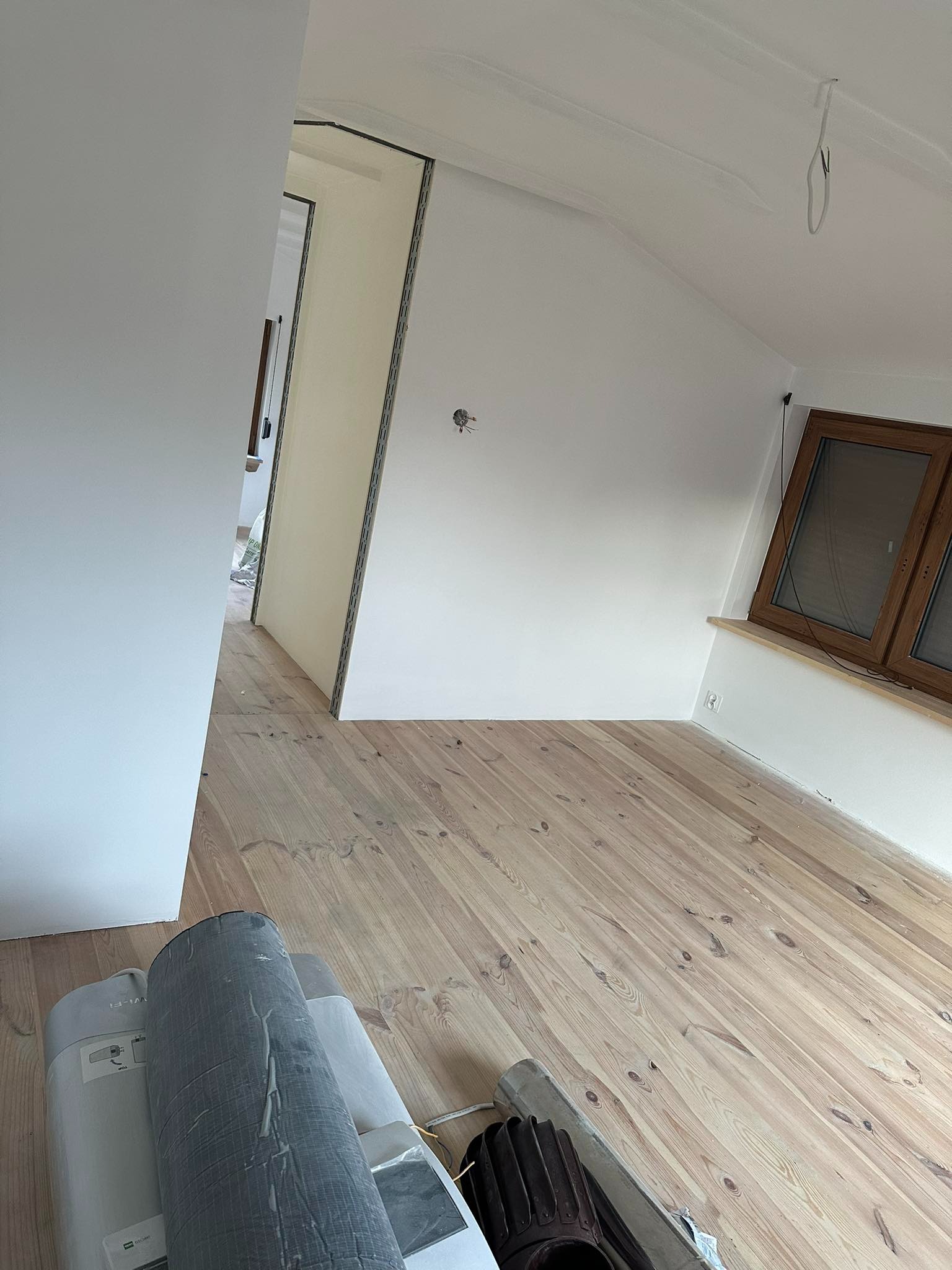 Poddasze po pracach wykończeniowych: świeżo pomalowane ściany w odcieniach bieli, drewniana podłoga, okno w drewnianej ramie, widoczne rolki materiałów budowlanych w rogu pomieszczenia.