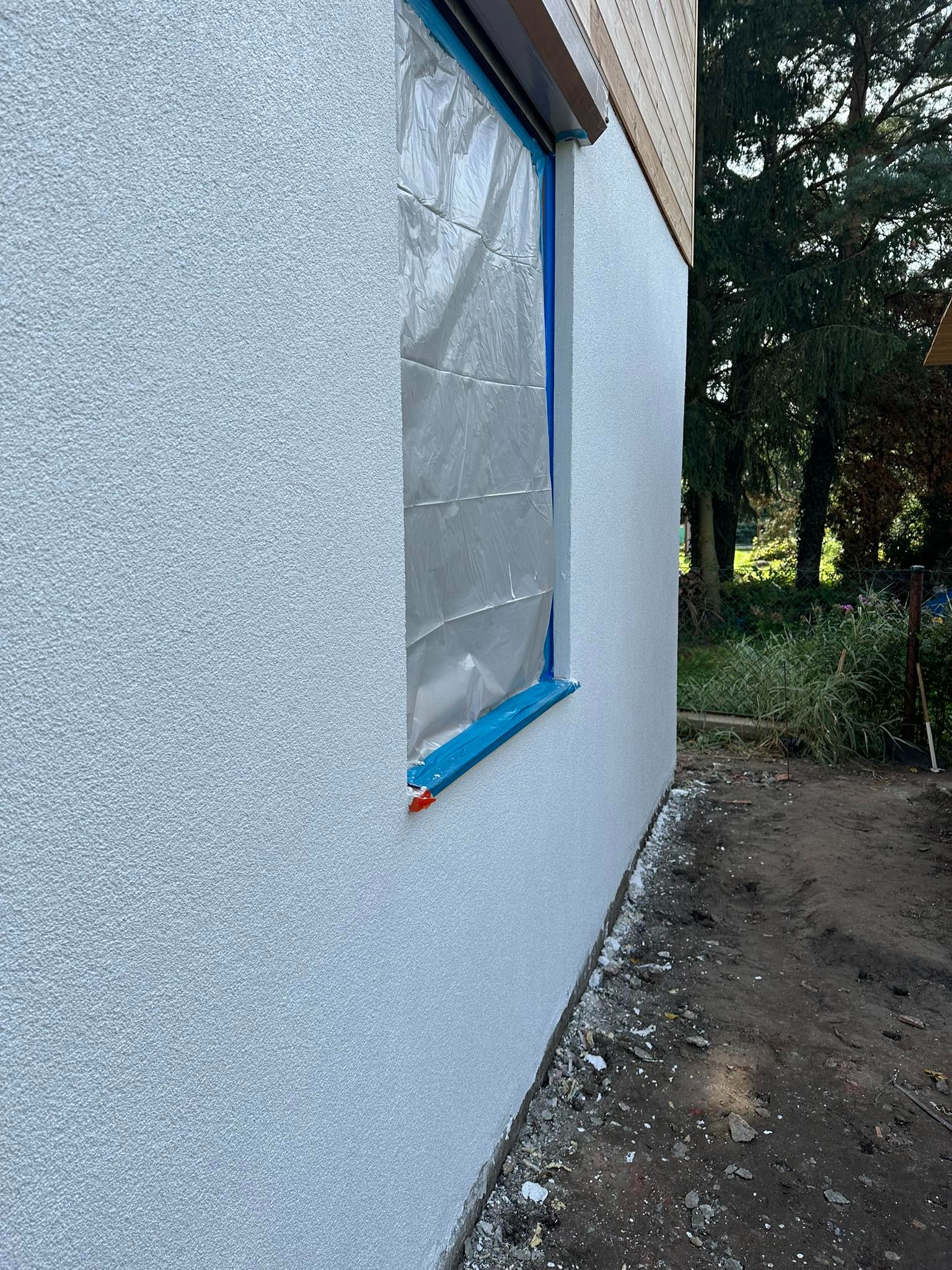 Ściana zewnętrzna budynku z nową, jasną strukturą. Okno zabezpieczone folią i taśmą malarską. Fragment drewnianej elewacji na górze. Grunt przy ścianie częściowo uprzątnięty, widoczna ziemia.
