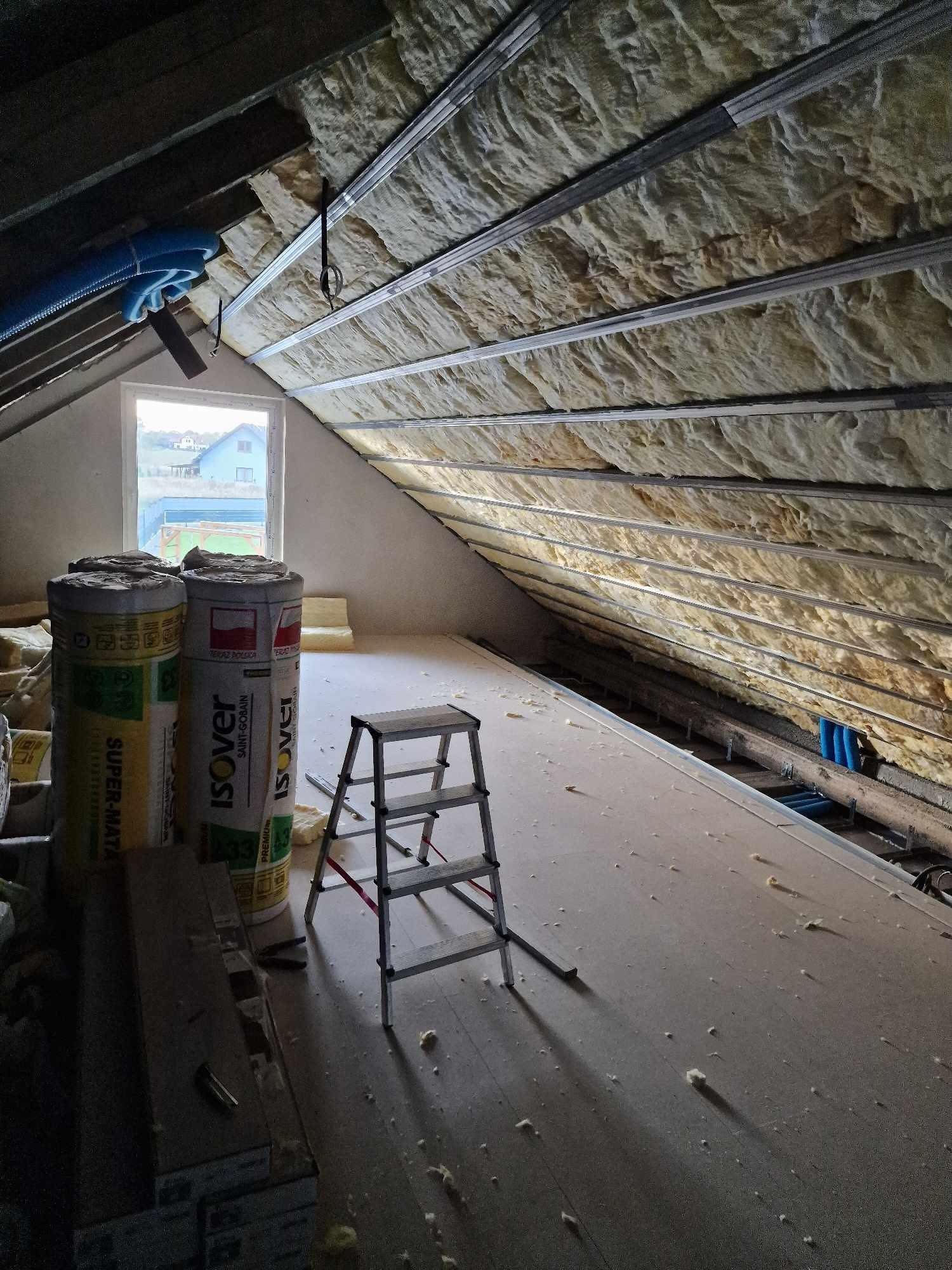 Poddasze w trakcie ocieplania wełną mineralną Isover, widoczne profile metalowe, drabina aluminiowa, rolki wełny mineralnej i okno dachowe z widokiem na domy.