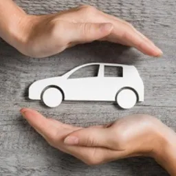 Dłonie formujące ochronny gest nad papierową sylwetką samochodu na drewnianym biurku, obok widoczny smartfon.