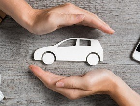 Dłonie formujące ochronny gest nad papierową sylwetką samochodu na drewnianym biurku, obok widoczny smartfon.