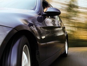 Czarny samochód w ruchu, widok z boku, rozmyte tło drzew, dynamiczne ujęcie z niskiej perspektywy.