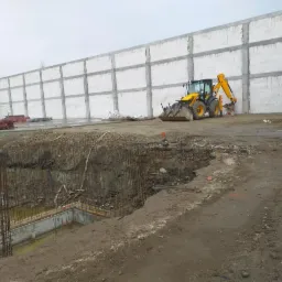 Żółta koparko-ładowarka JCB stoi obok wykopu zbrojonego stalą, w tle surowy budynek z bloczków betonowych, widoczne elementy szalunków.