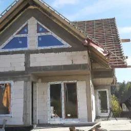 Budowa domu murowanego w Nowym Targu: surowy stan z oknami o nietypowym tr&oacute;jkątnym kształcie i częściowo wykonaną konstrukcją dachu, widoczne materiały izolacyjne.