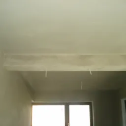 Wnętrze w trakcie prac wykończeniowych: świeżo otynkowane ściany i sufit w jasnym odcieniu, widoczne wystające przewody z sufitu, w tle okno z jasnym światłem.