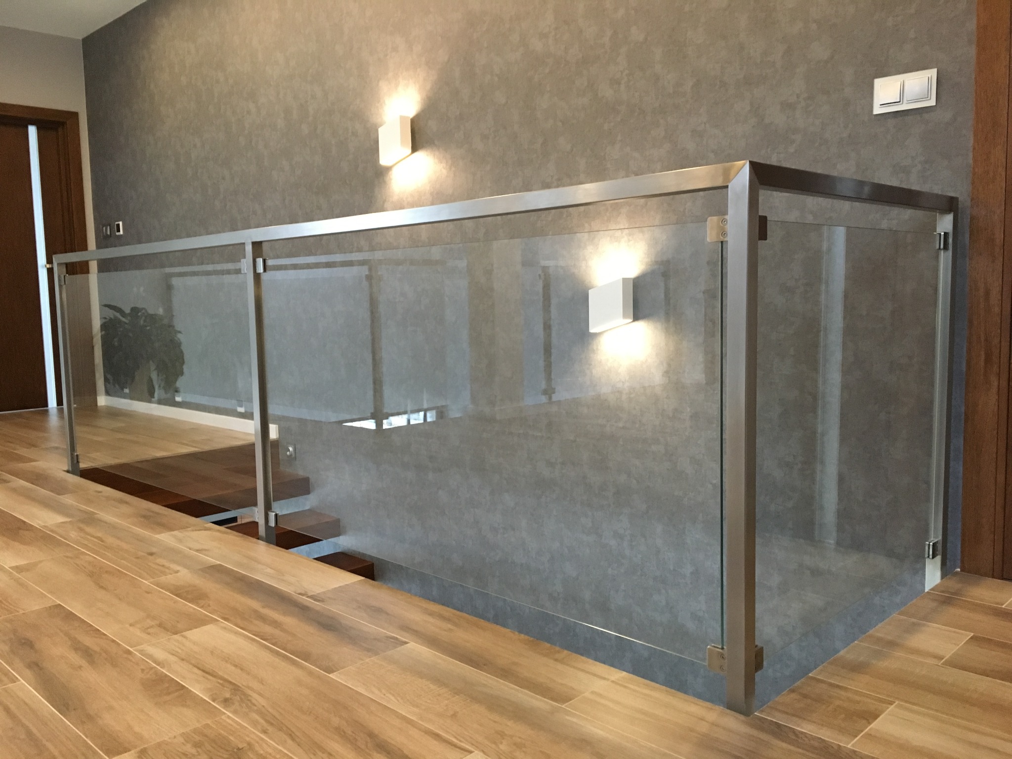 Nowoczesne wnętrze z szklaną balustradą i metalową ramą, oświetlone kinkietami na szarej ścianie, widok na drewniane schody i podłogę z paneli imitujących drewno.