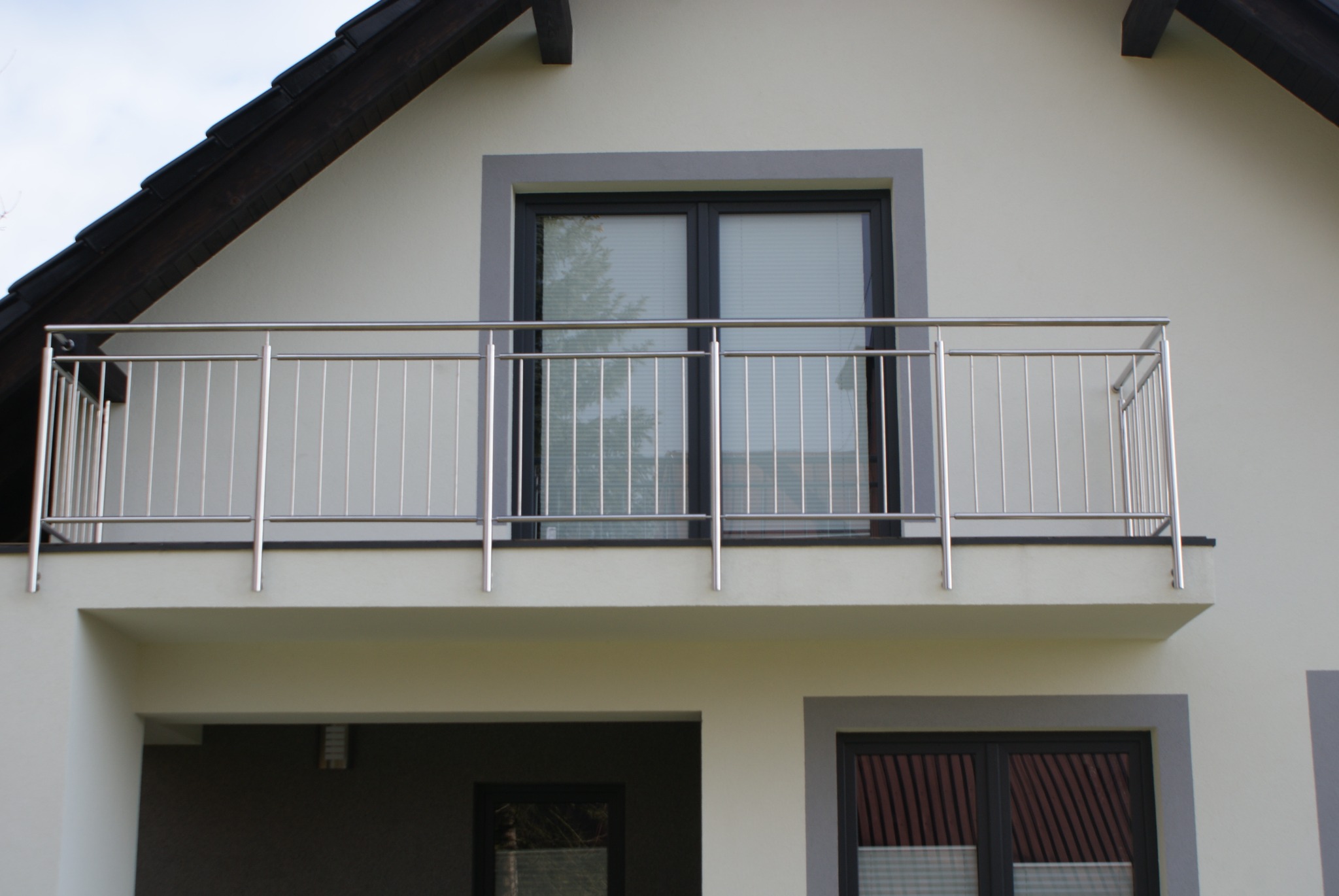 Balkon z prostą, nowoczesną balustradą ze stali nierdzewnej, widok z dołu na jasną elewację budynku z oknami w ciemnych ramach.