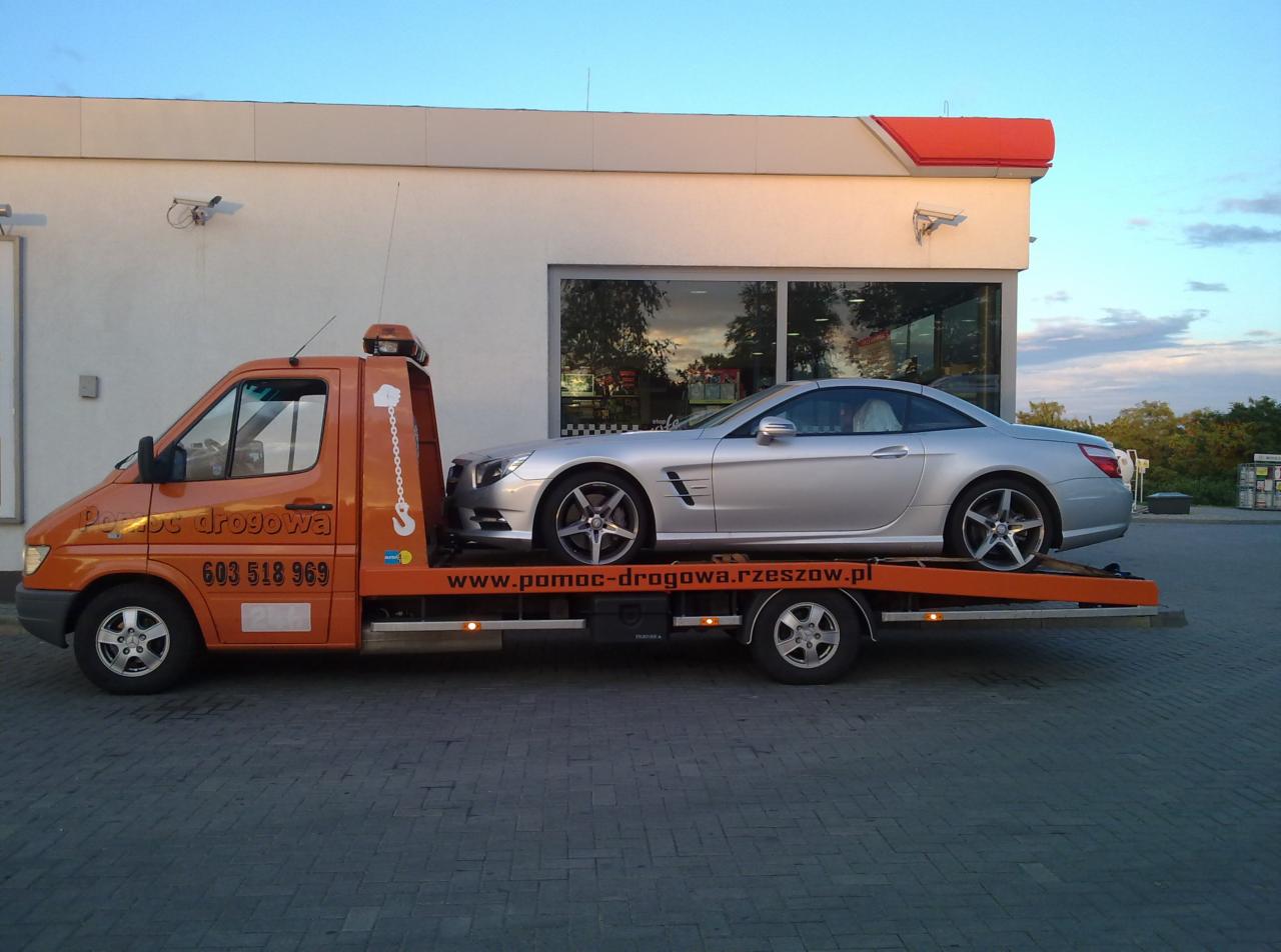 Pomarańczowa laweta Mercedes-Benz Sprinter z srebrnym Mercedesem SL na platformie, zaparkowana przed budynkiem z dużymi oknami i kamerami monitoringu.