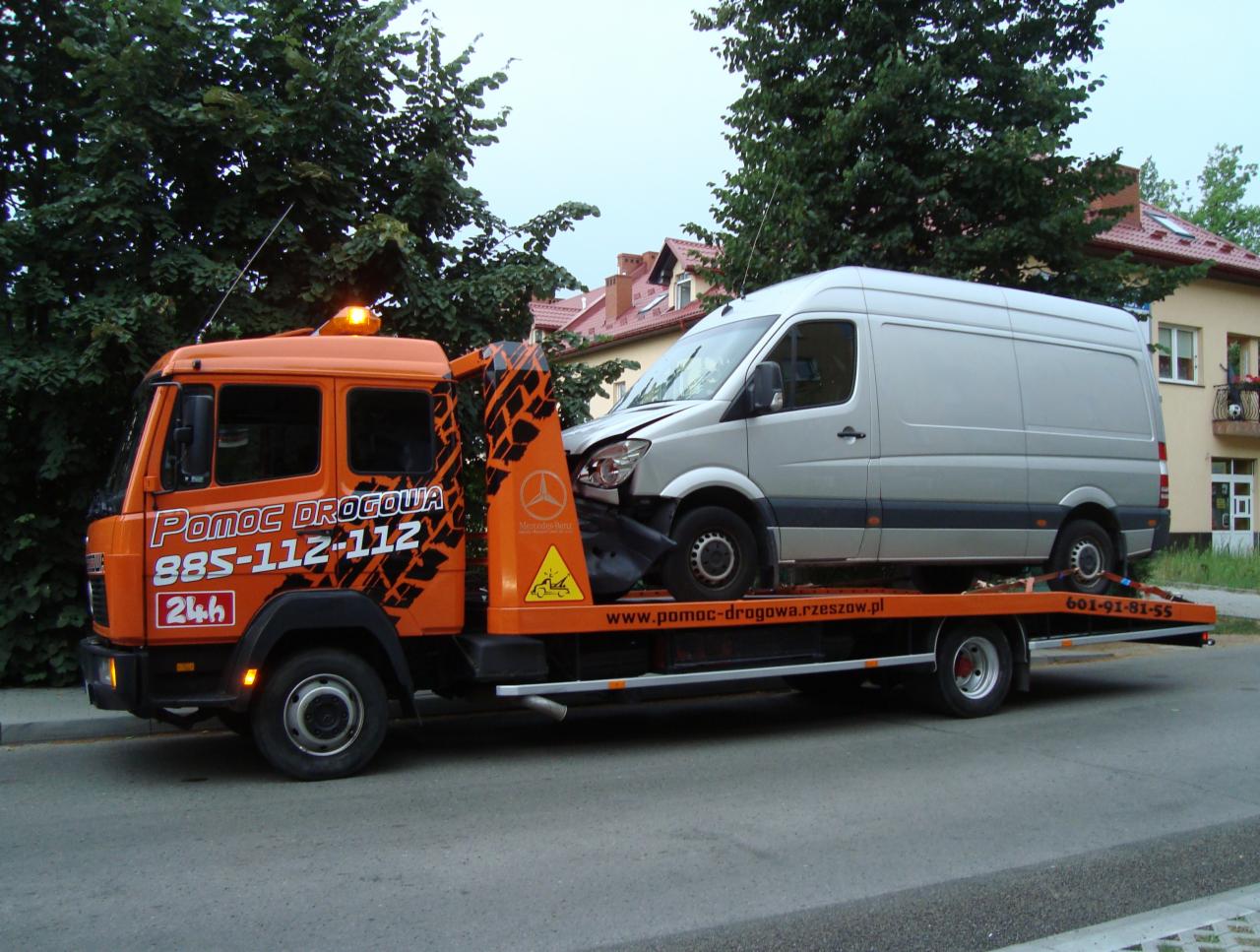 Pomarańczowa laweta z uszkodzonym srebrnym busem Mercedes na platformie, widoczne logo Mercedes-Benz i numer telefonu alarmowego na lawetcie, tło z zielenią i budynkami.