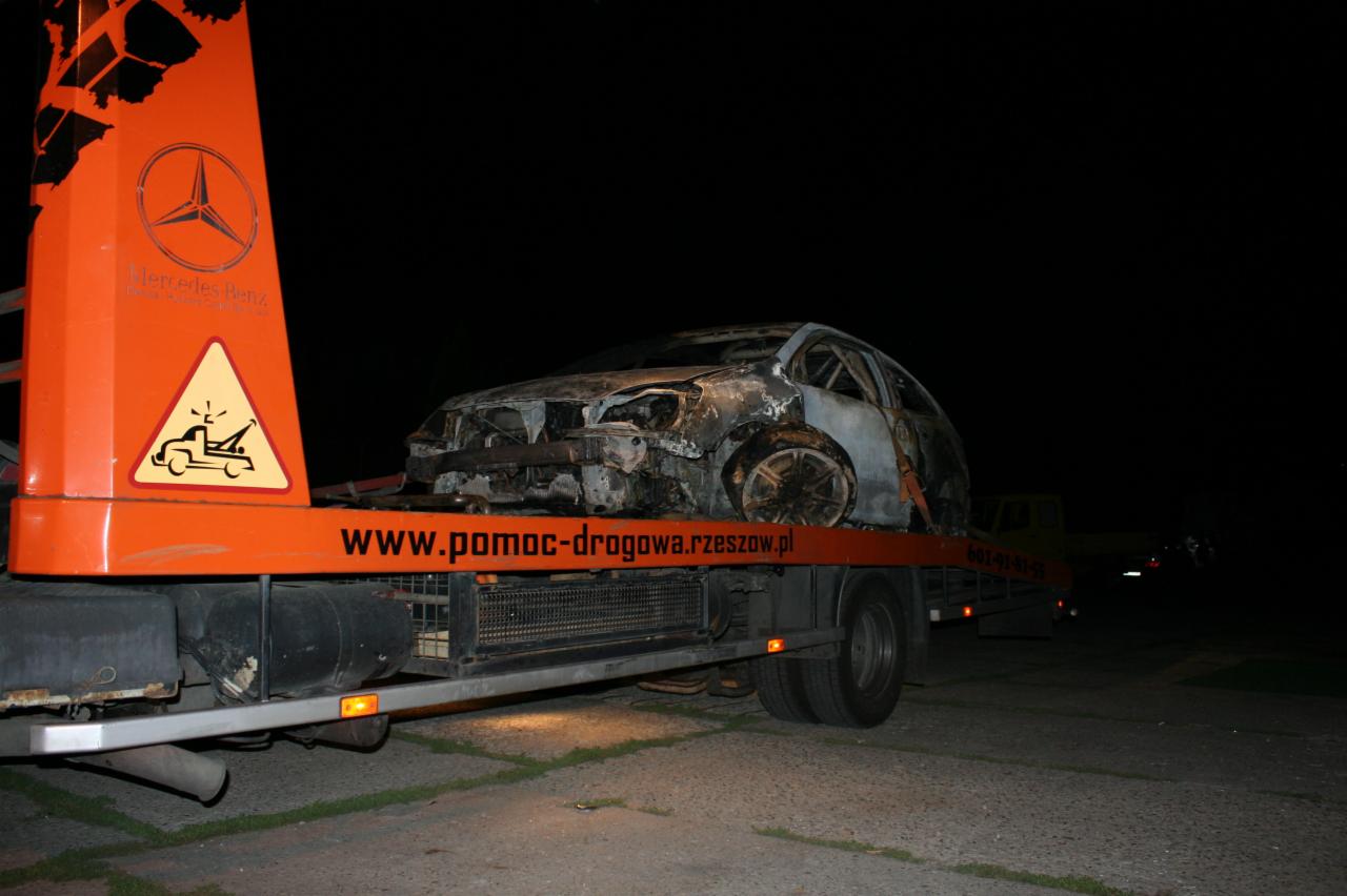 Spalony wrak samochodu osobowego przewożony na pomarańczowej lawecie pomocy drogowej www.pomoc-drogowa.rzeszow.pl w nocnych warunkach.