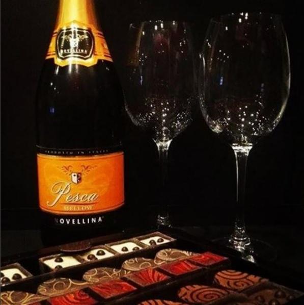 Butelka włoskiego wina musującego Pesca Mellow Novellina, dwa kieliszki do wina i pudełko ekskluzywnych czekoladek na czarnym tle.
