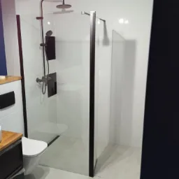 Nowoczesna kabina prysznicowa typu walk-in z czarnymi profilami, deszczownicą i baterią w kolorze czarnym, białe płytki na ścianach i podłodze, oświetlenie punktowe na suficie.