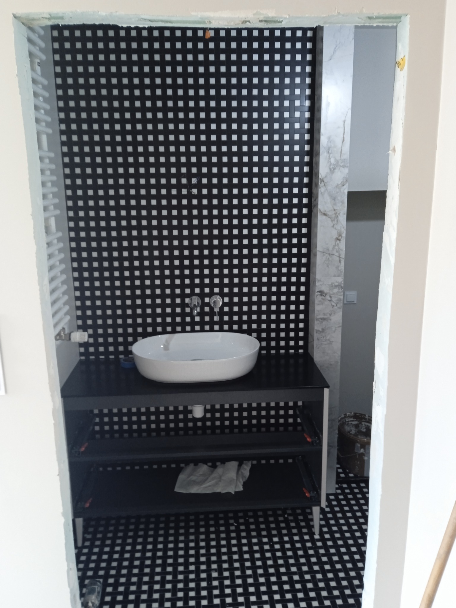 Łazienka w trakcie wykańczania: biała umywalka nablatowa, czarna szafka z półkami, ściana i podłoga w czarno-białą kratę, widoczne ślady prac wykończeniowych na ścianach.