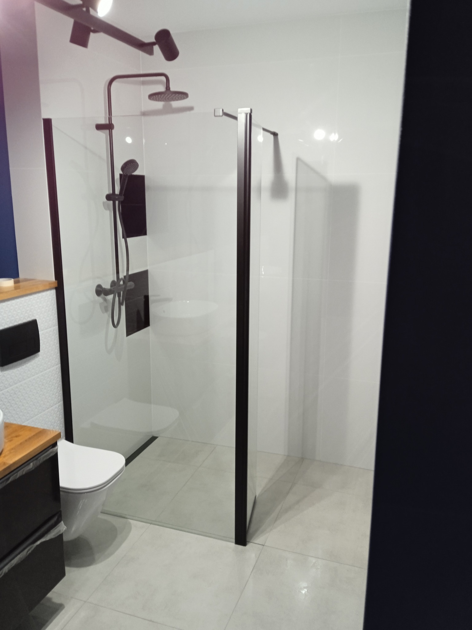 Nowoczesna kabina prysznicowa typu walk-in z czarnymi profilami, deszczownicą i baterią w kolorze czarnym, białe płytki na ścianach i podłodze, oświetlenie punktowe na suficie.